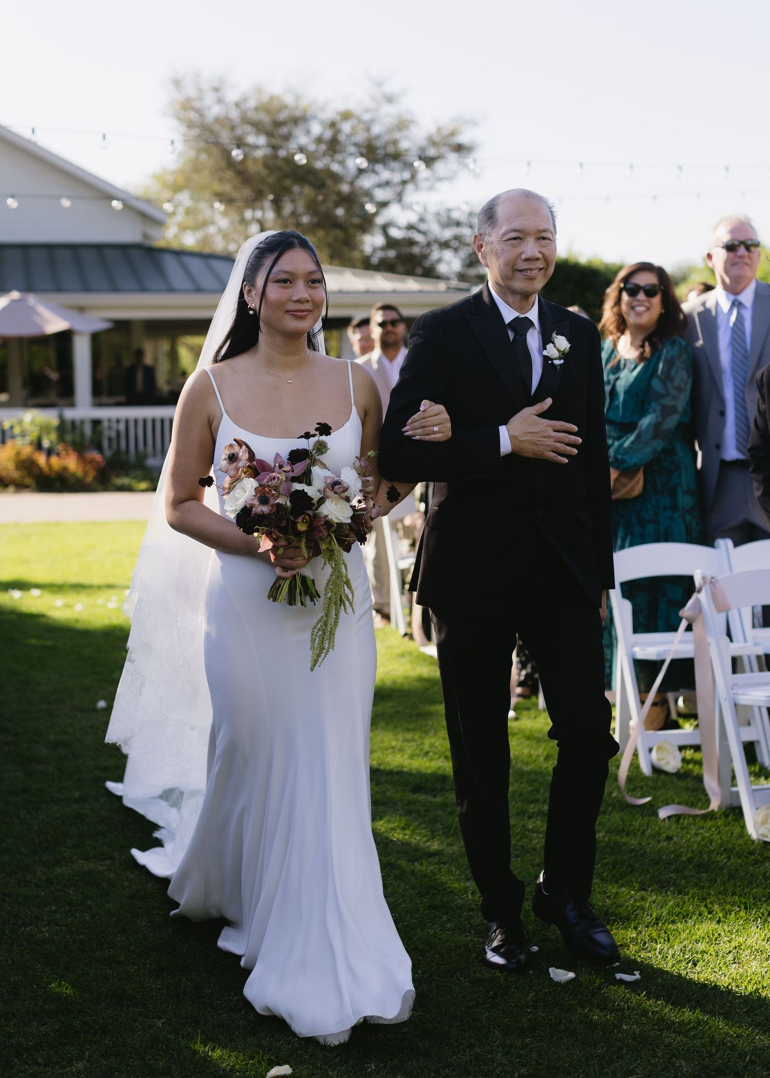 Wedding party walking down an outdoor aisle with white chairs and green grass.