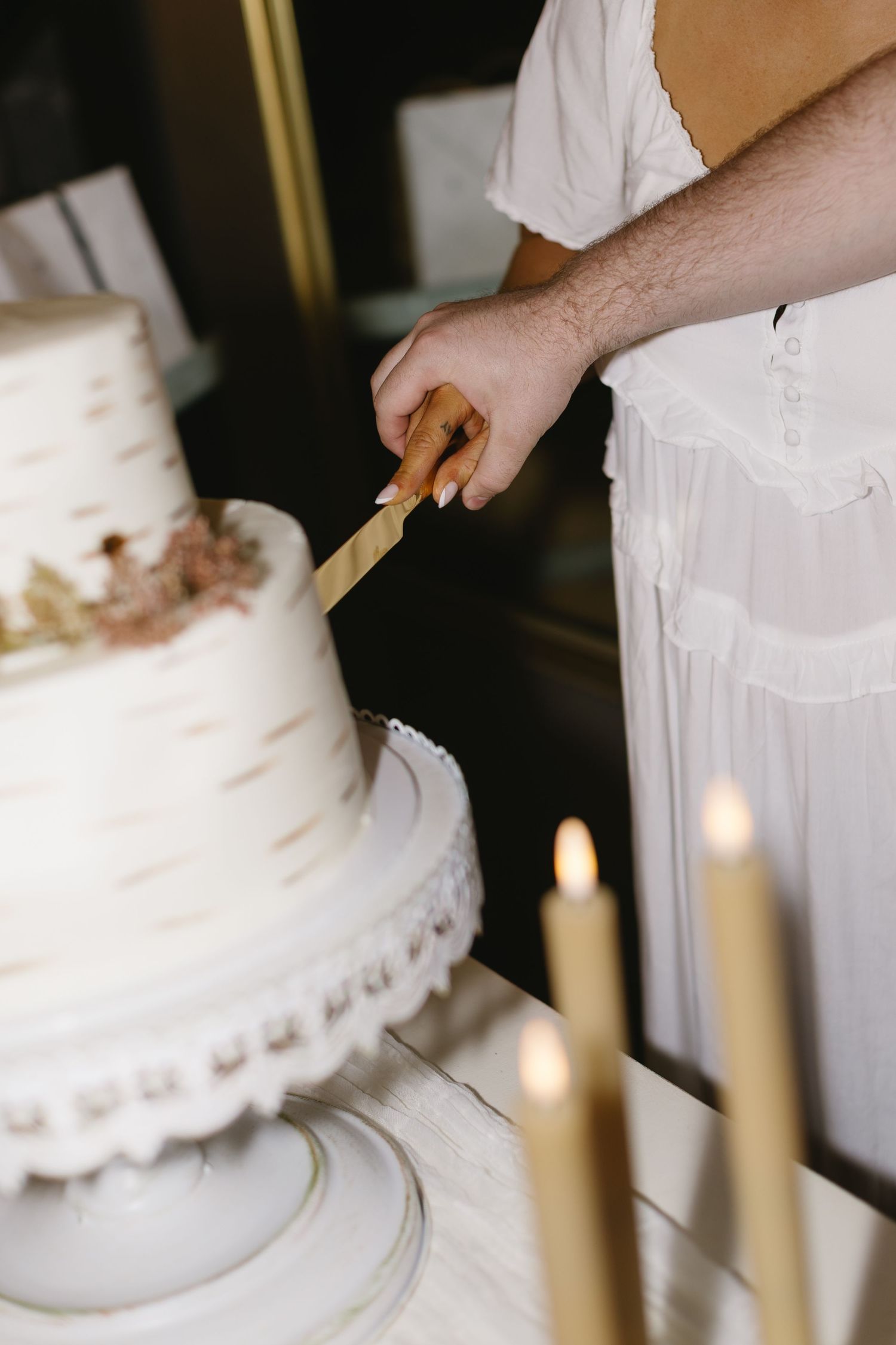 Hands cutting into a white wedding cake with lit candles in the foreground.