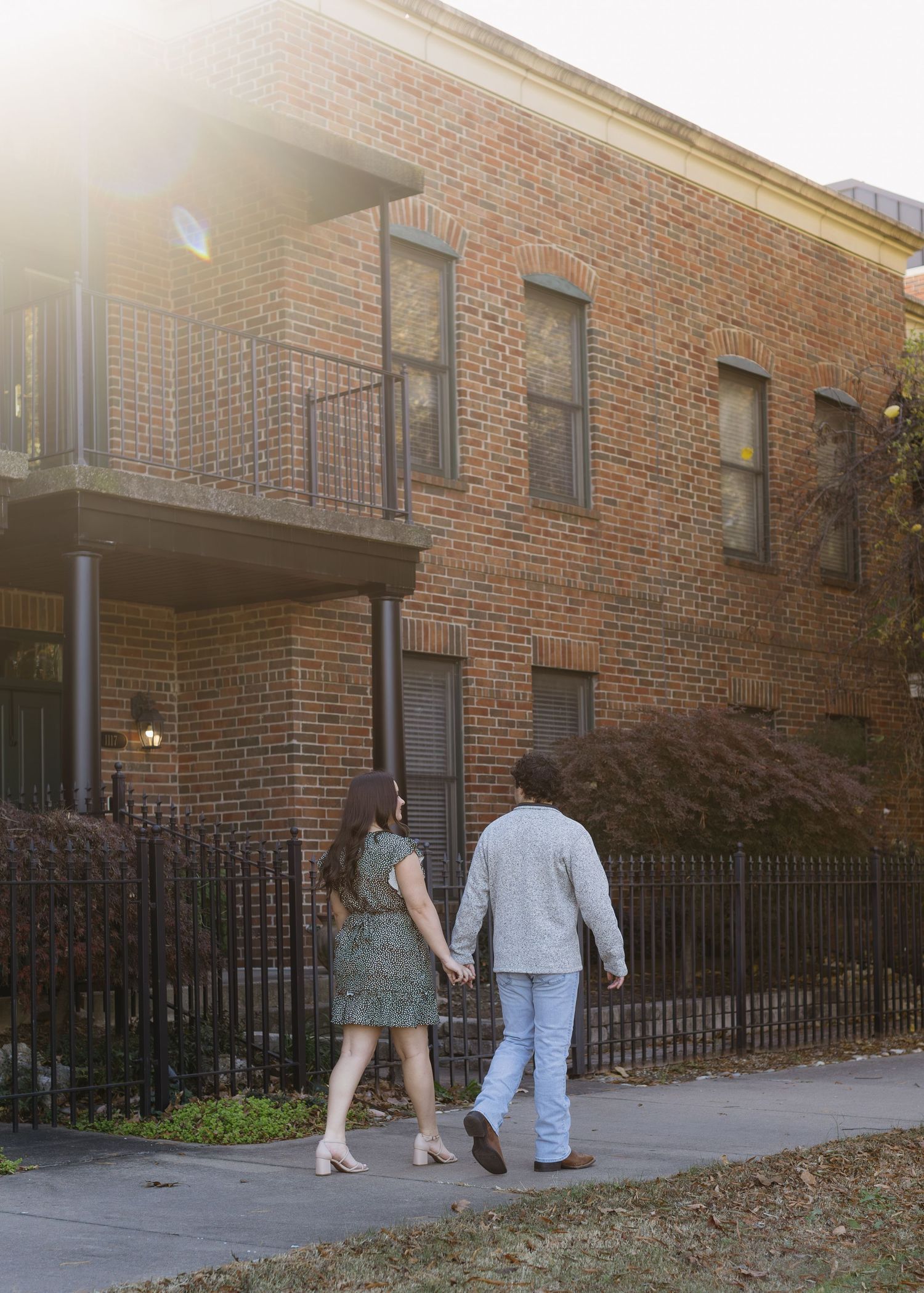 Two people walk along a brick building pathway with wrought iron fencing on an autumn day.