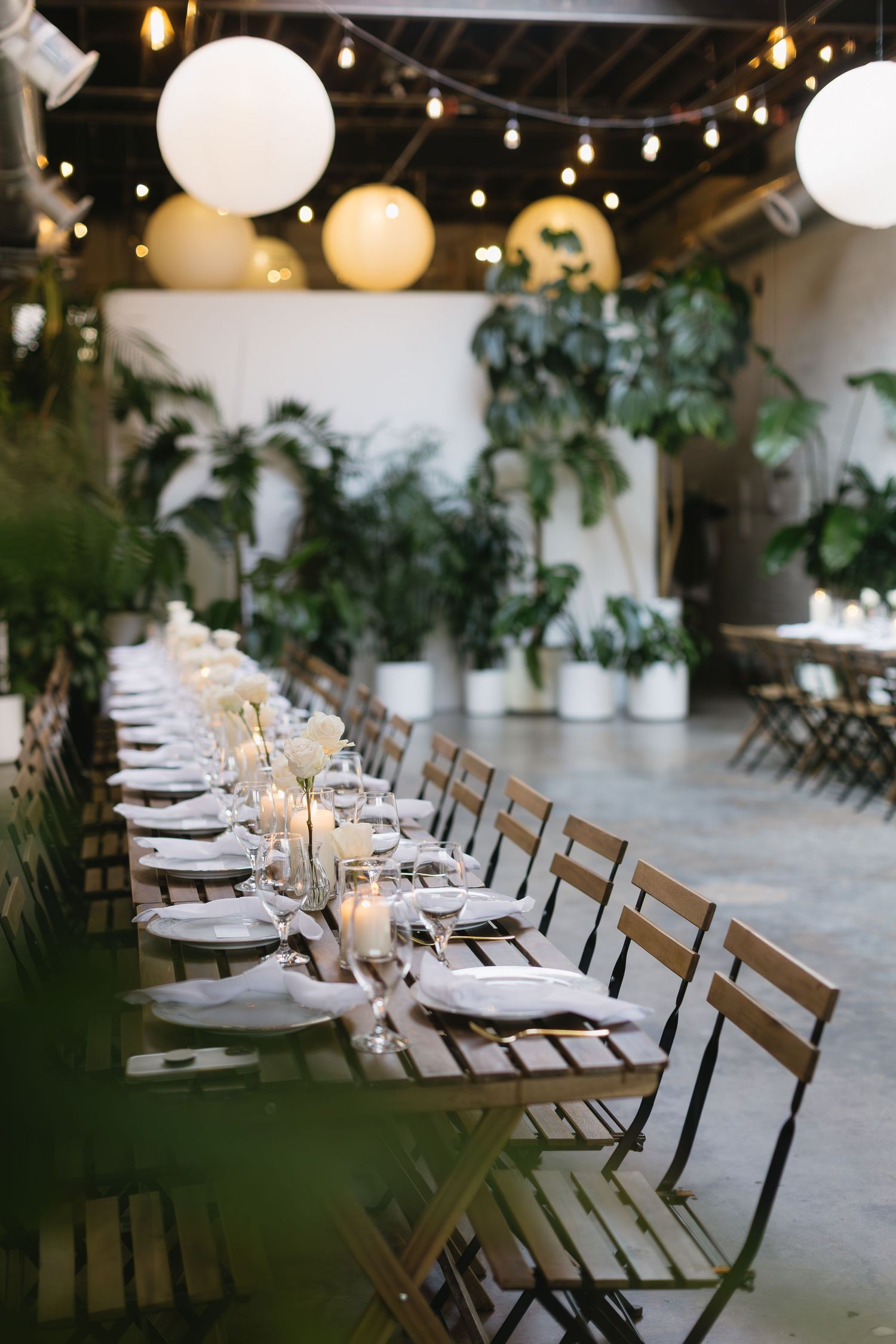 A rustic dining setup features wooden chairs and white place settings under warm string lights and paper lanterns.