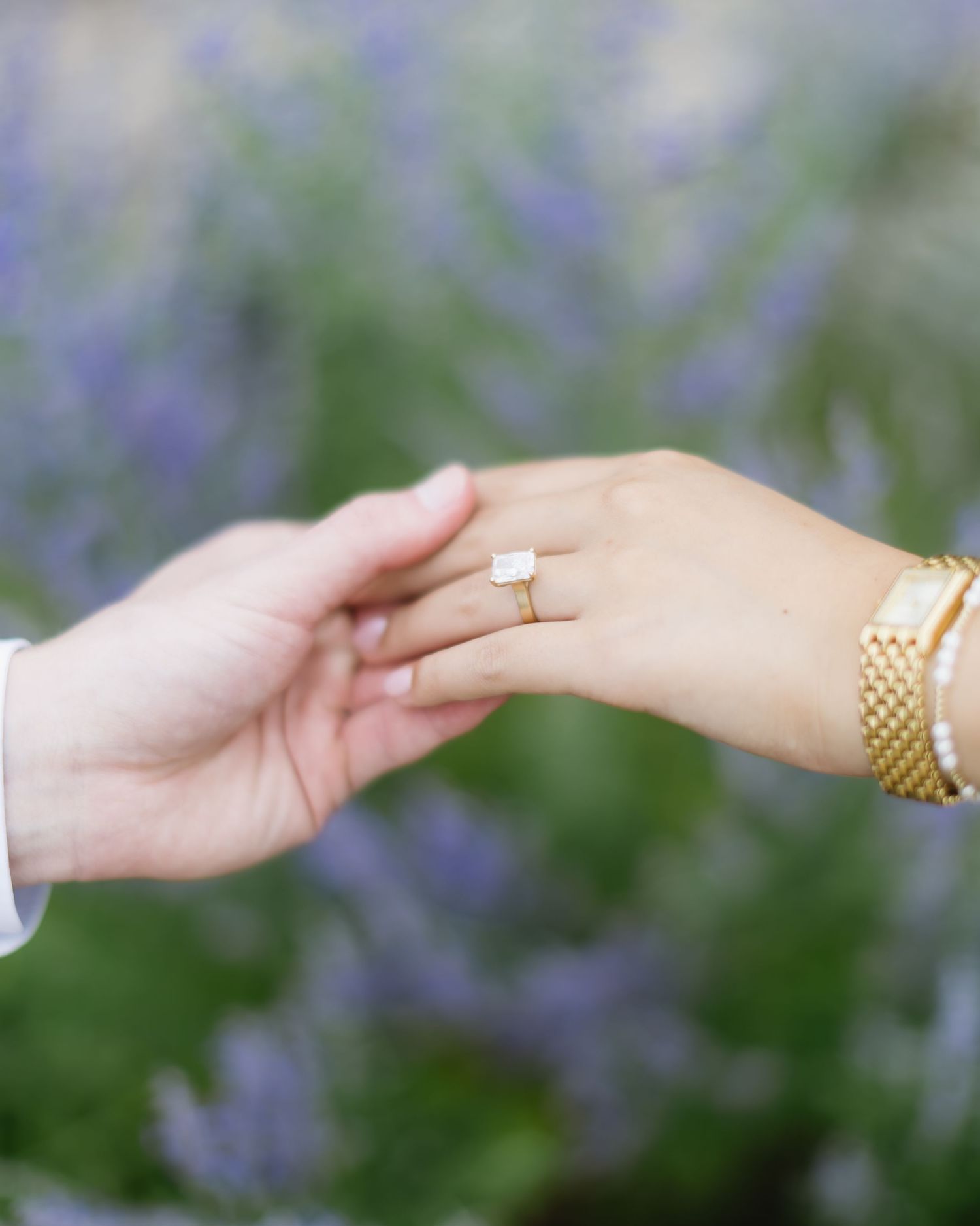 Close-up of hands reaching towards each other showing wedding rings and jewelry against a blurred floral background.