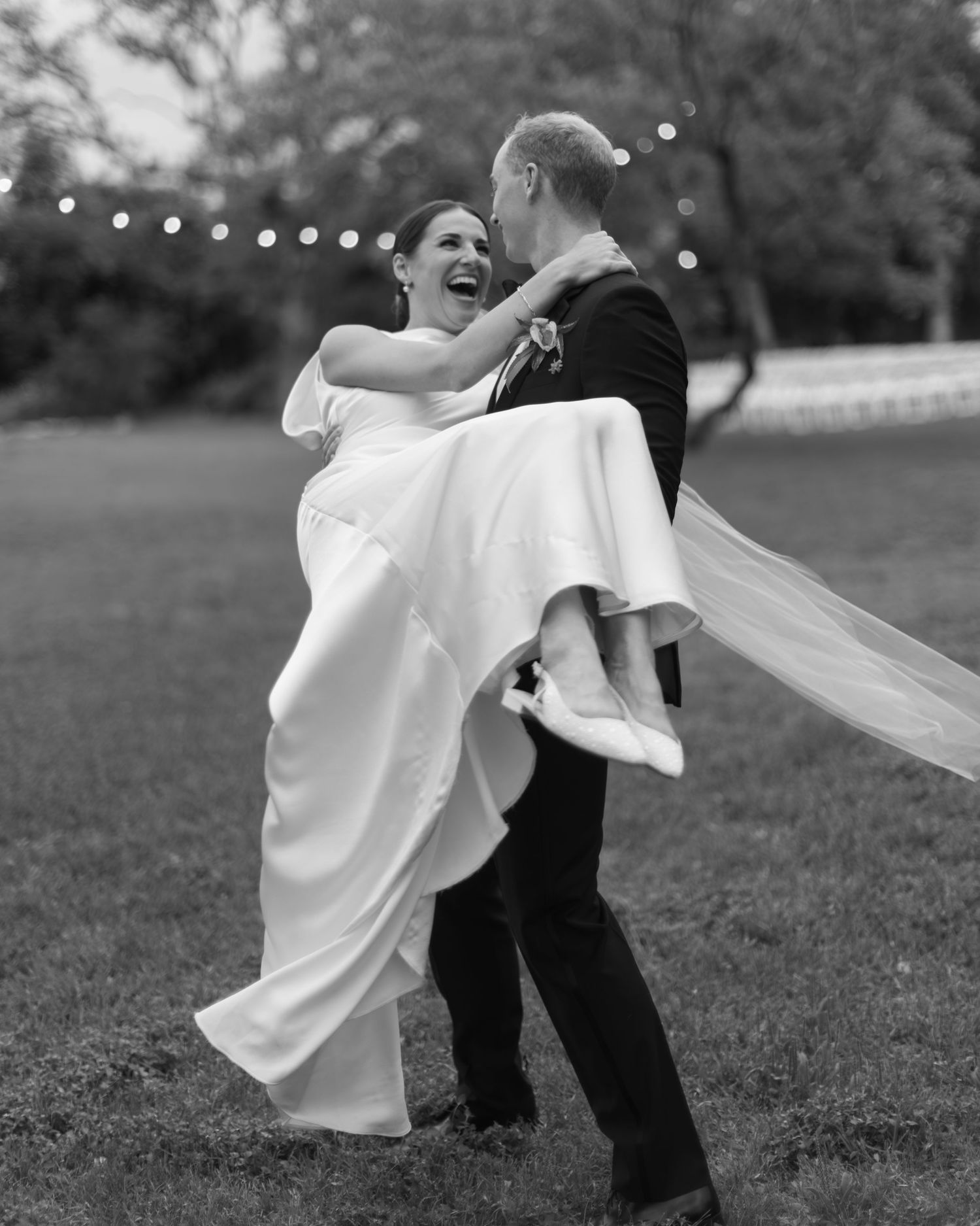 A black and white wedding photo of a joyful couple dancing outdoors with string lights in the background.