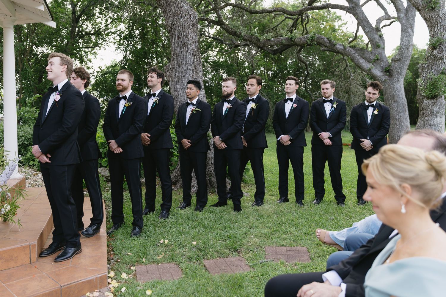 Wedding party lined up outdoors with groomsmen in black tuxedos.