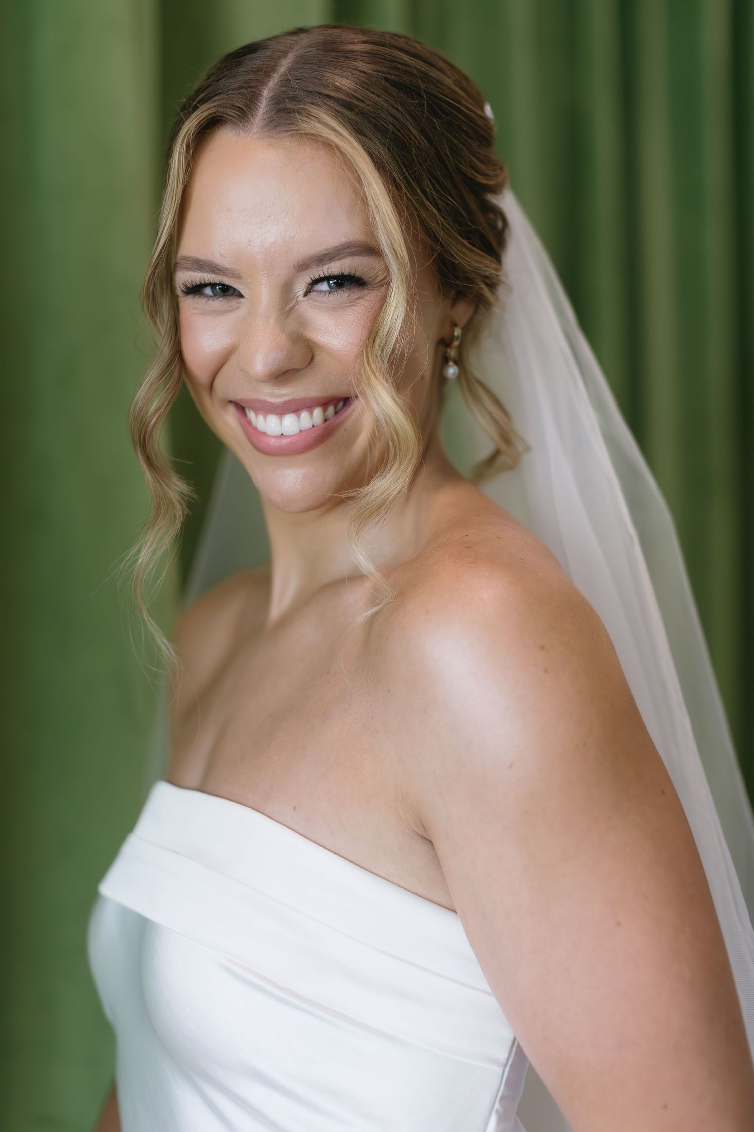 Bride in white strapless gown and veil smiles joyfully against a green drape background.