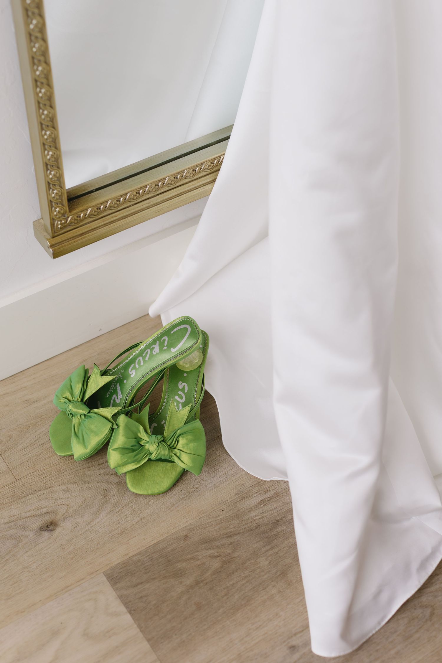 Green satin shoes sit on a wooden floor beside white curtains and gold-framed mirrors.