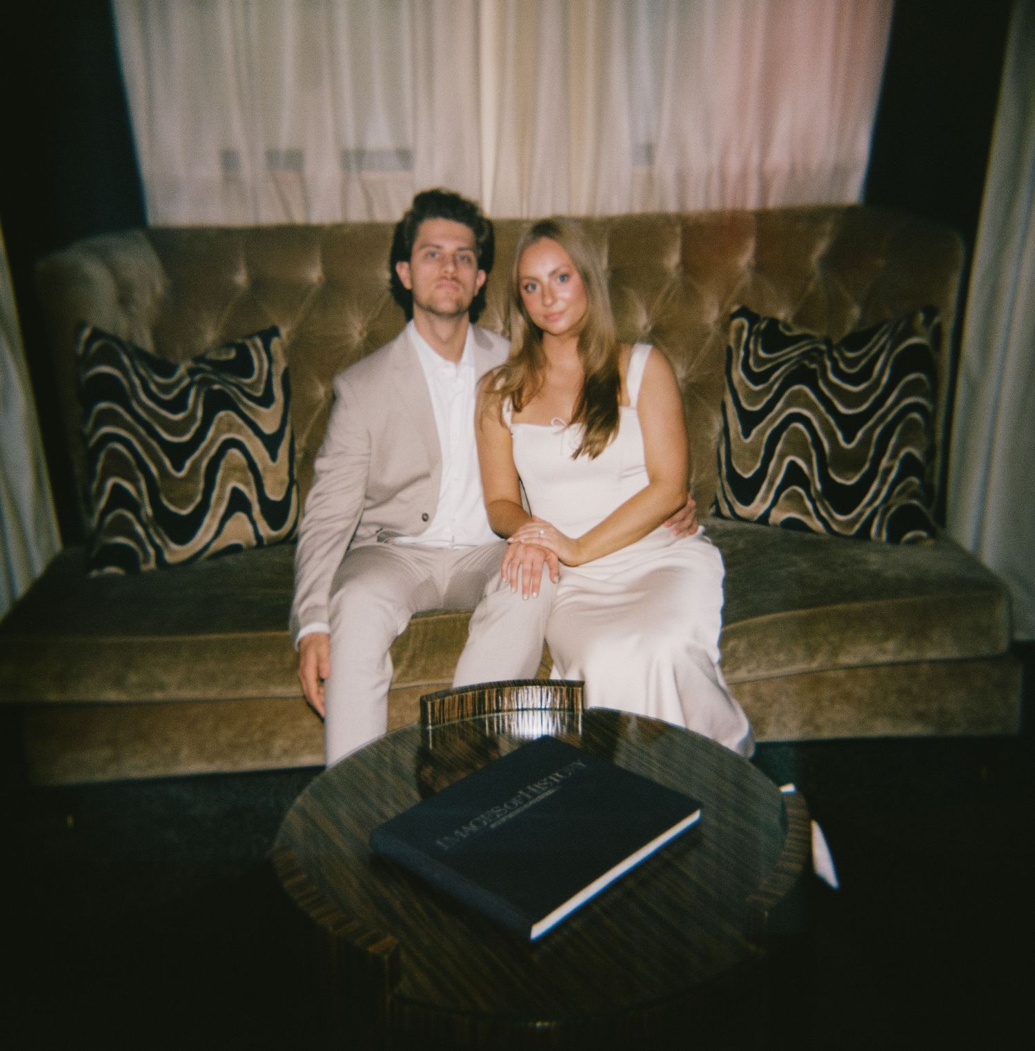 A couple in formal attire sits together on a dark patterned couch with a coffee table in front.