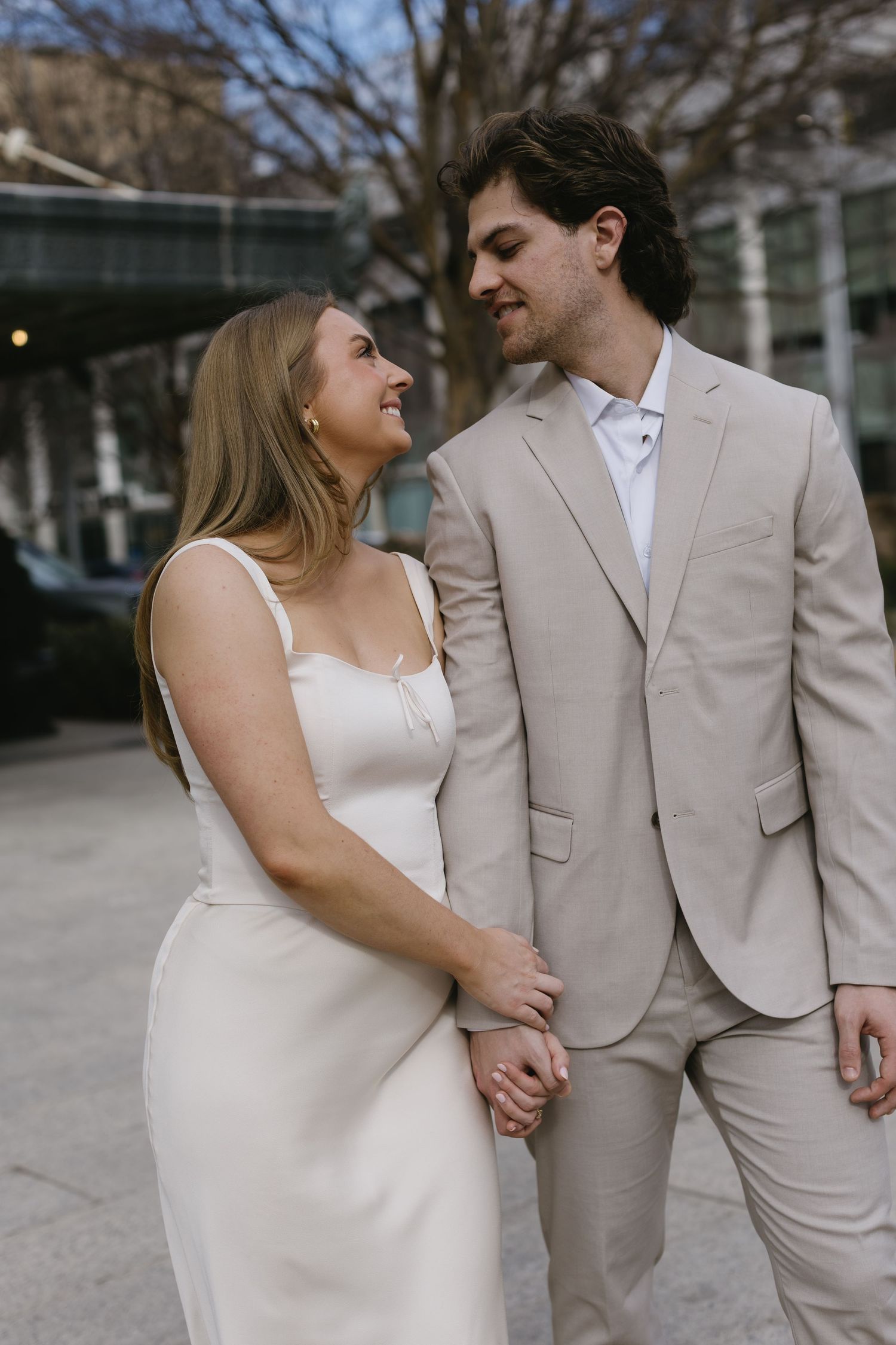 A couple in coordinated white dress and beige suit pose together outdoors.