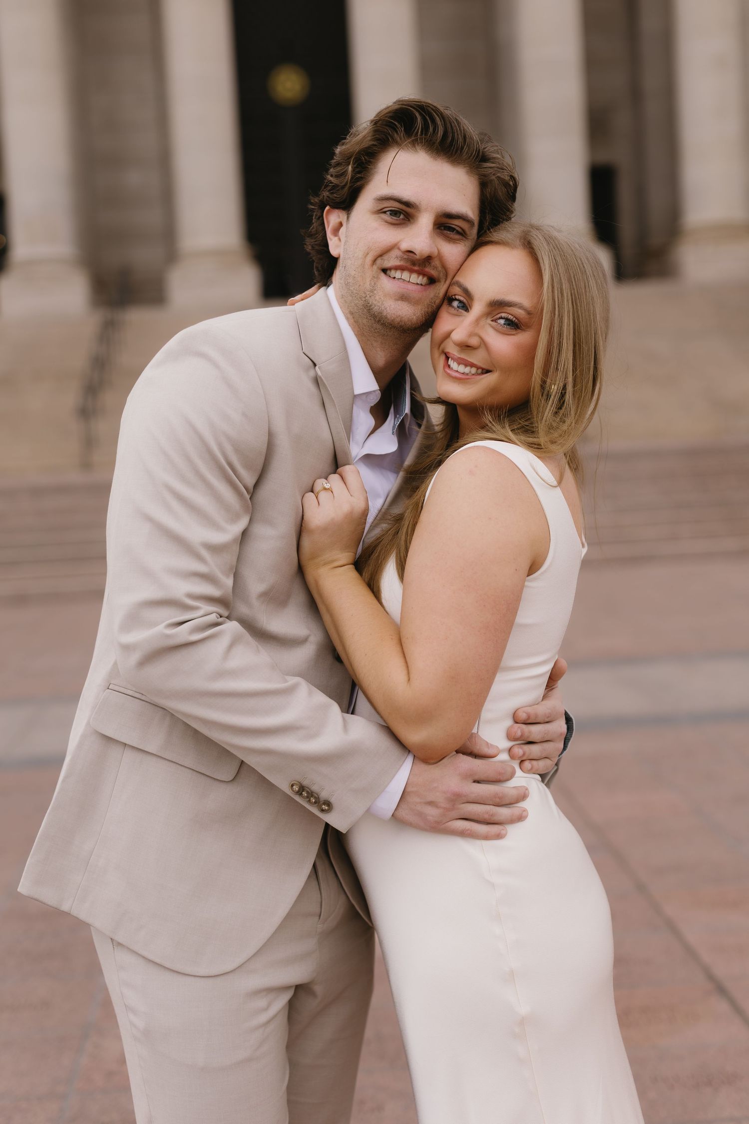 A joyful couple in light colored formal wear shares a laugh while embracing outdoors.