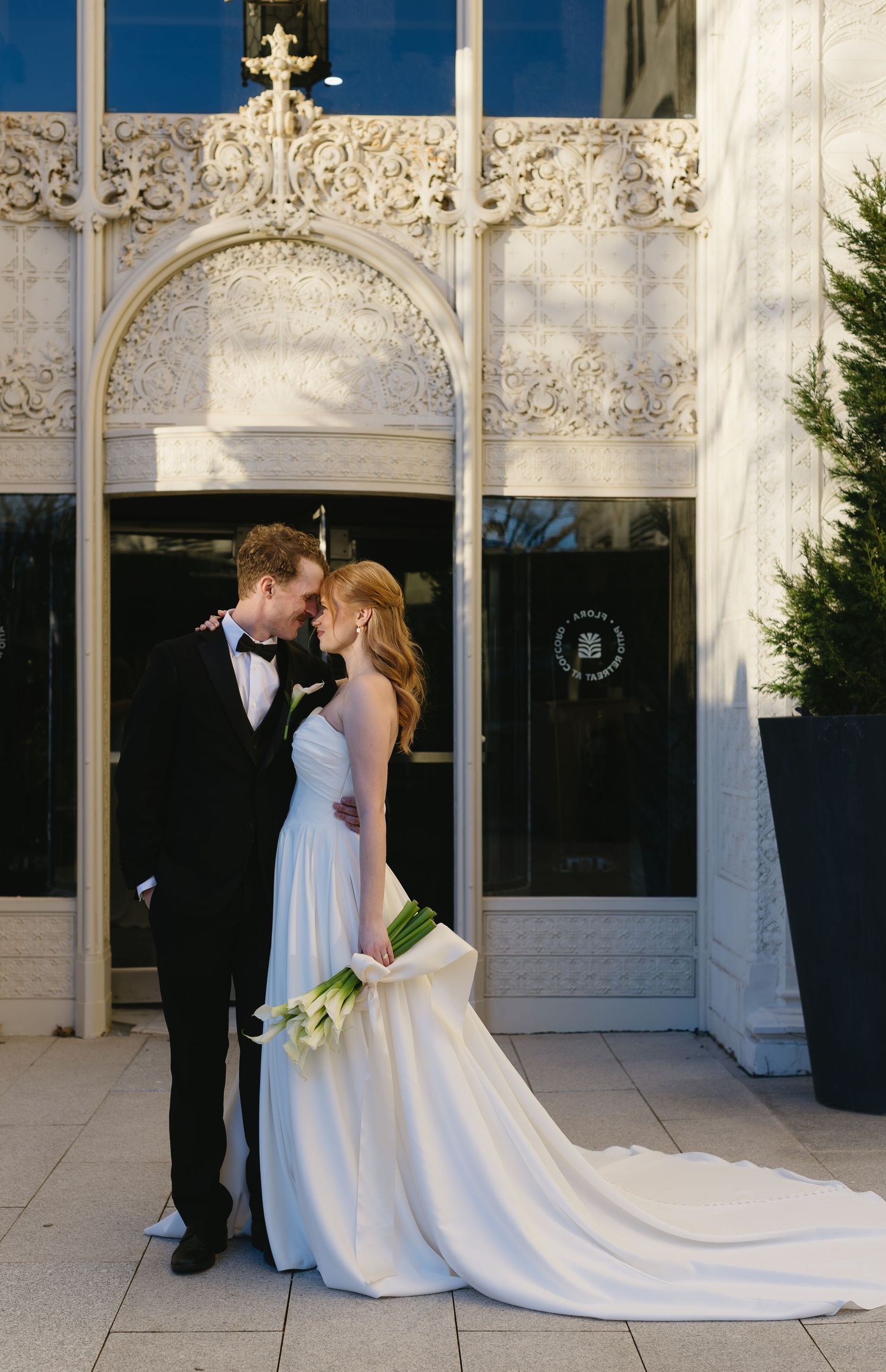 Elegant wedding couple poses outside ornate white stone building with classical architecture.