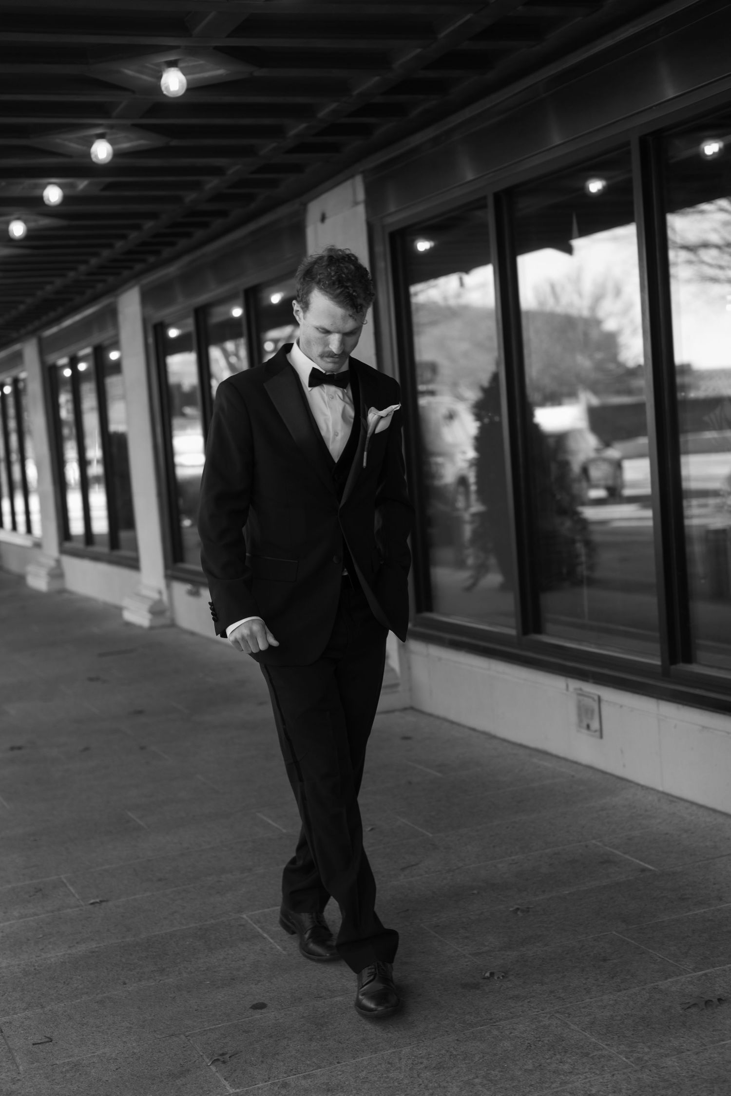 Black and white image showing a formal sequence of walking feet on a covered walkway with overhead lights.