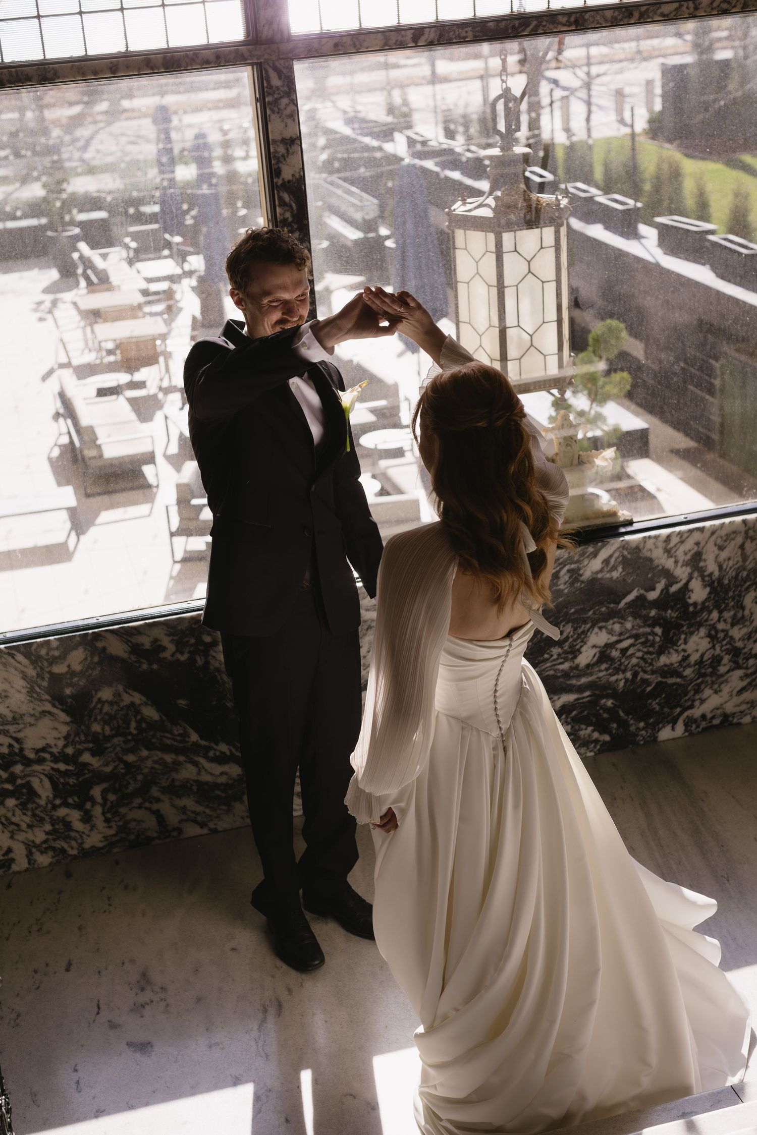 A couple shares an intimate moment in formal wedding attire against a city backdrop through floor-to-ceiling windows.