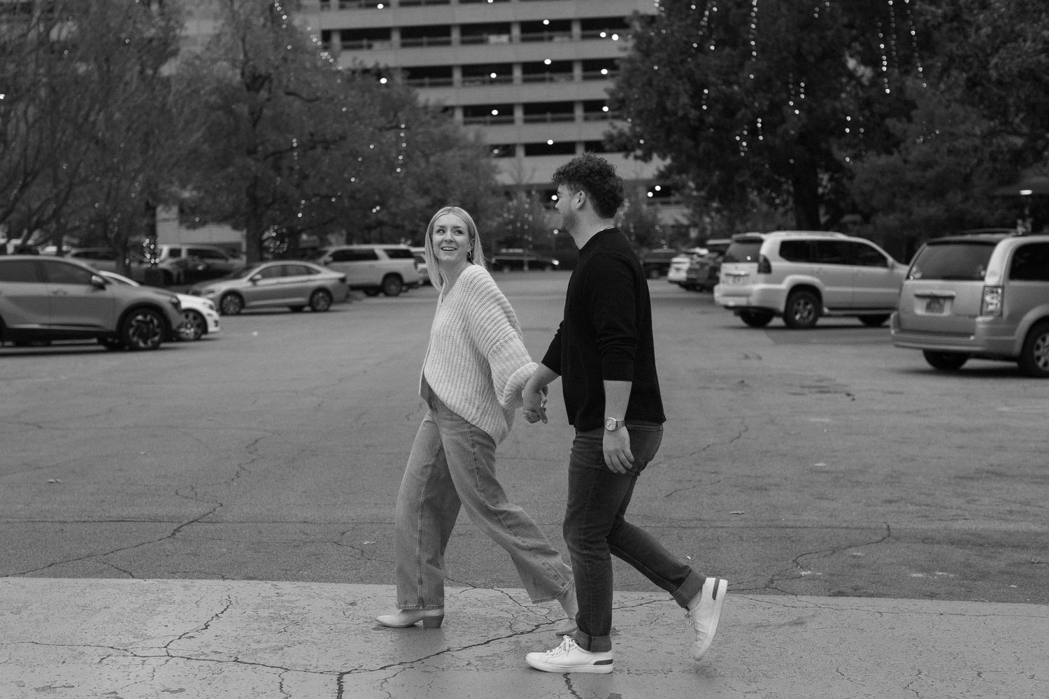 A black and white photo of a couple walking playfully through a parking lot.