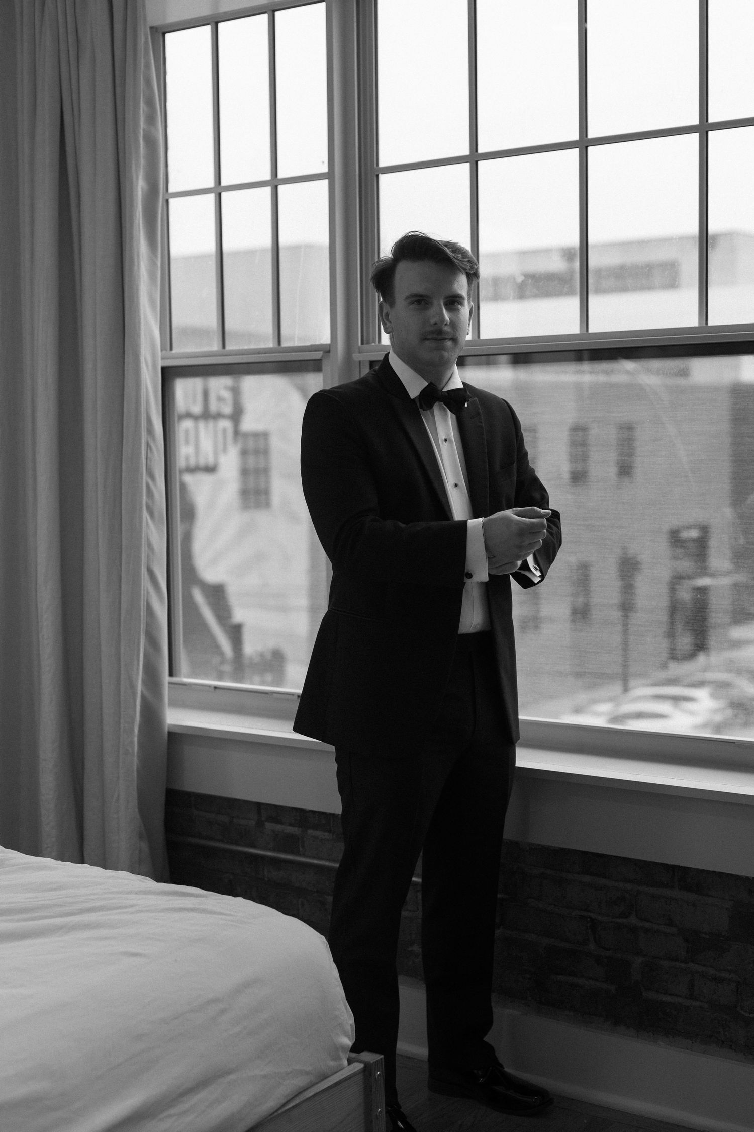 A black and white photo shows a person in a dark suit standing by a window in an empty room.