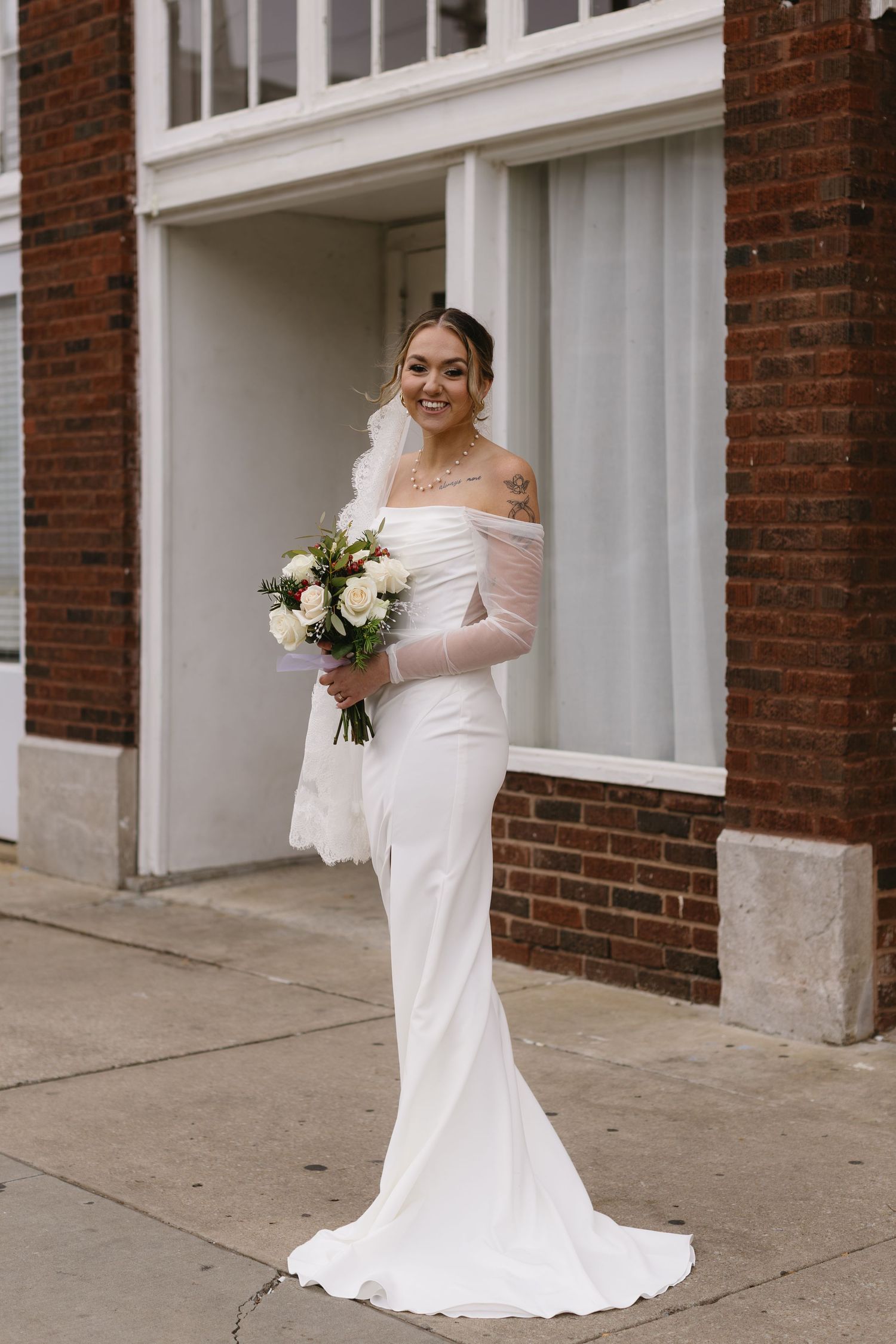 Bride in fitted white gown standing against brick building with white windows.