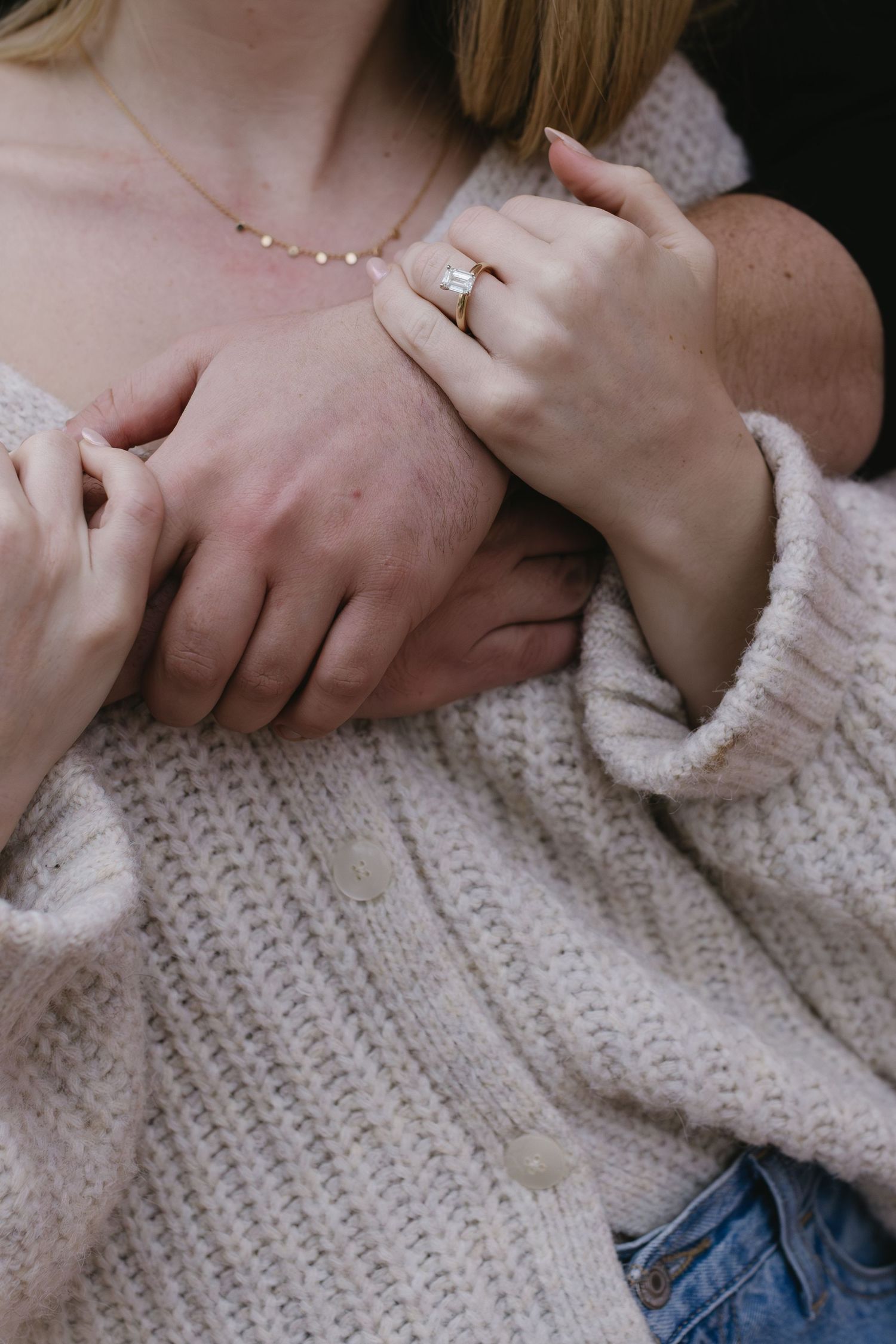 Close-up of hands wearing an engagement ring with a delicate necklace and cream sweater.