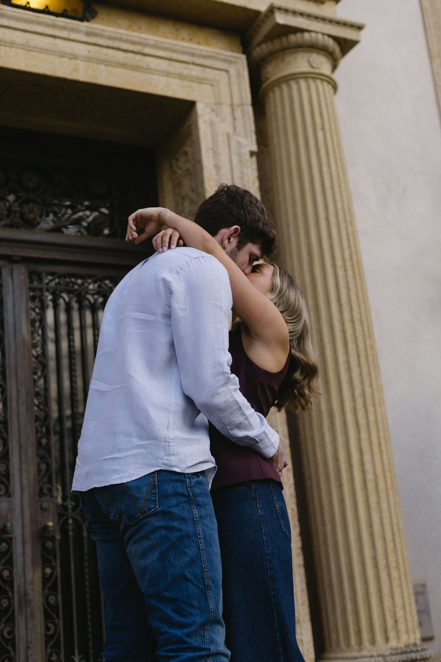 A couple shares an intimate embrace near a classical column and doorway.