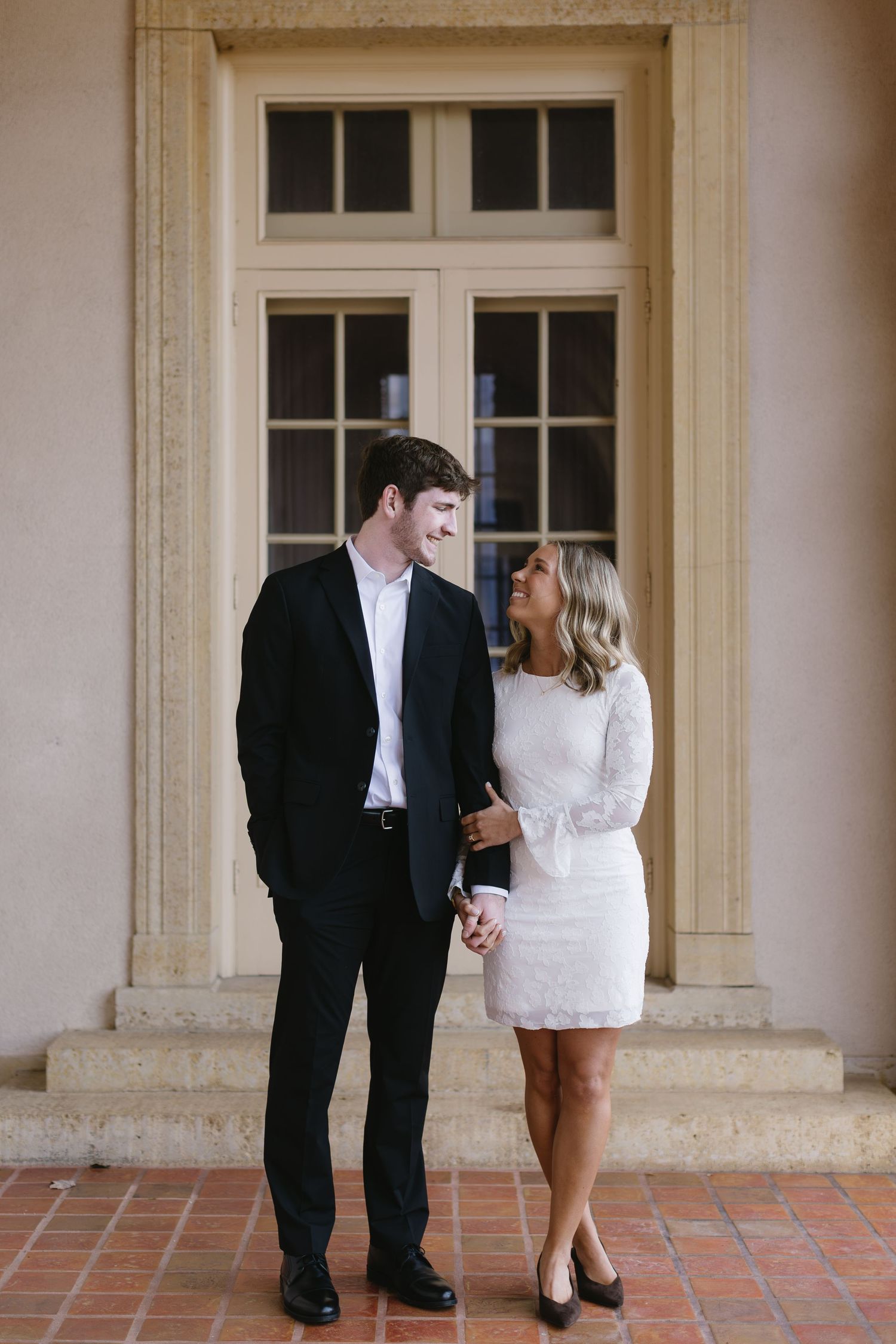 A romantic engagement photo series showing a couple in formal attire on a tiled patio near elegant windows.