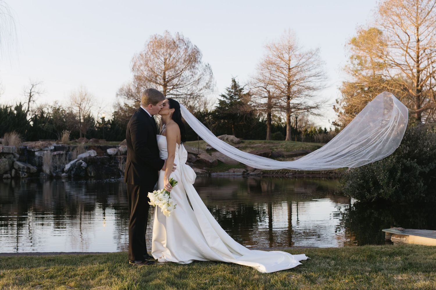 Wedding dress and veil dramatically flowing in breeze by lakeside setting.