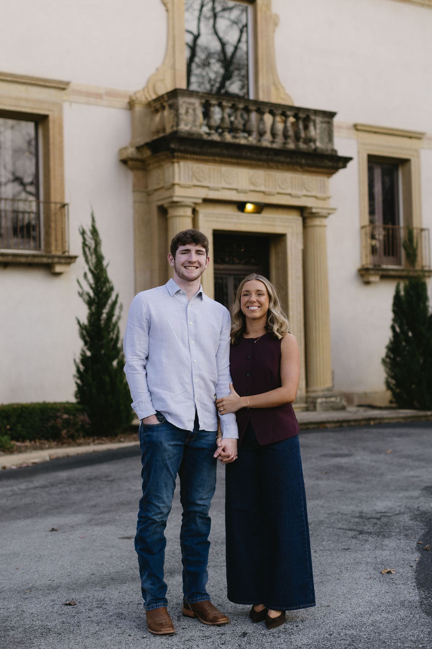 A couple poses together against a cream-colored building with Spanish architectural details.