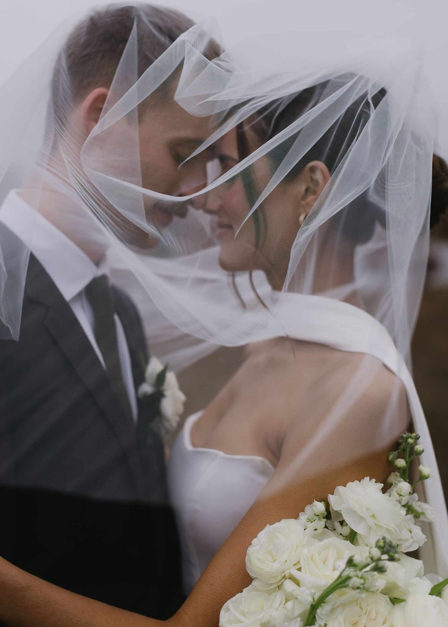 Wedding couple shares intimate moment under bridal veil with white roses in foreground.