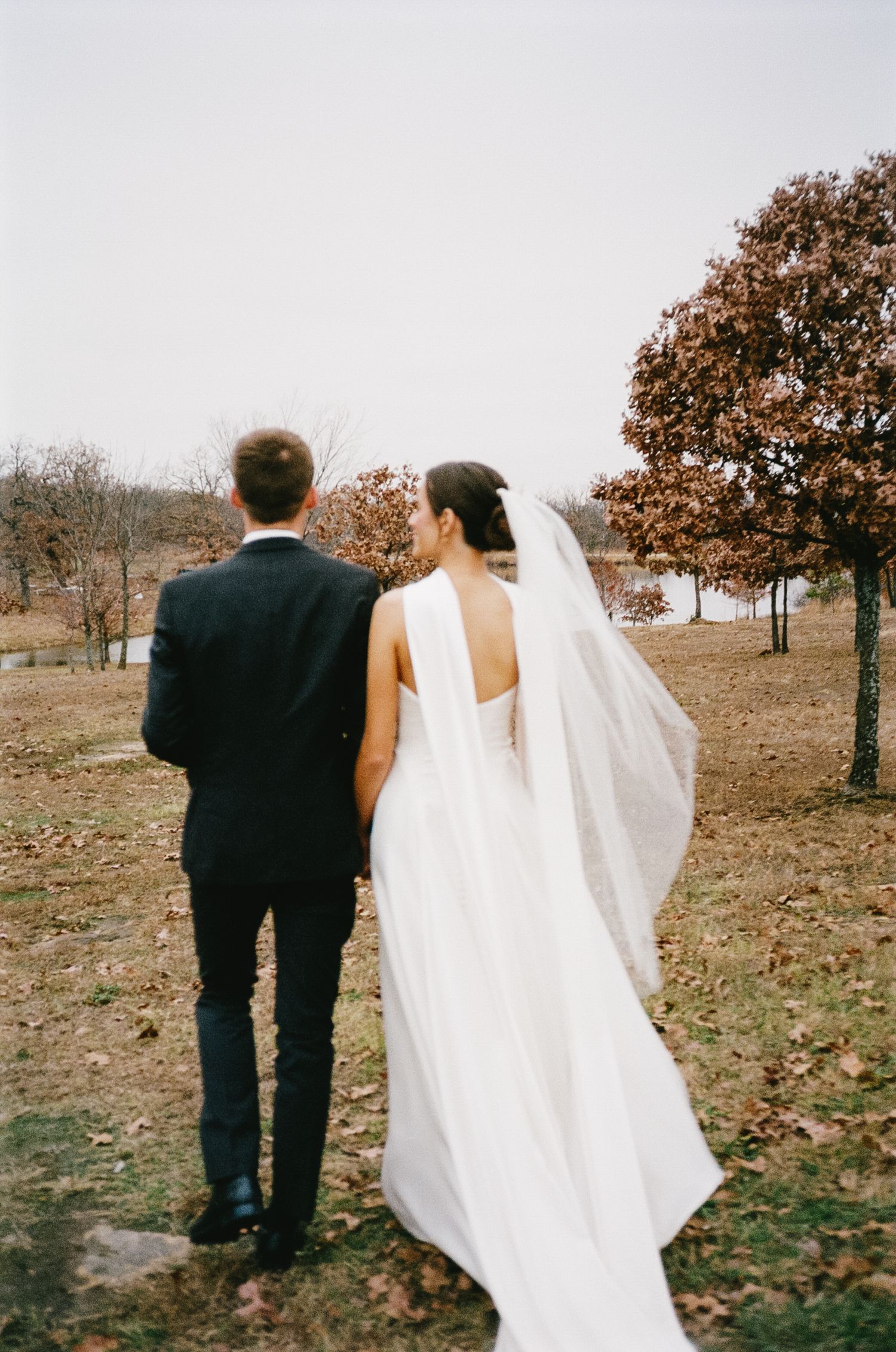 Bride and groom walking together through autumn trees in formal attire.