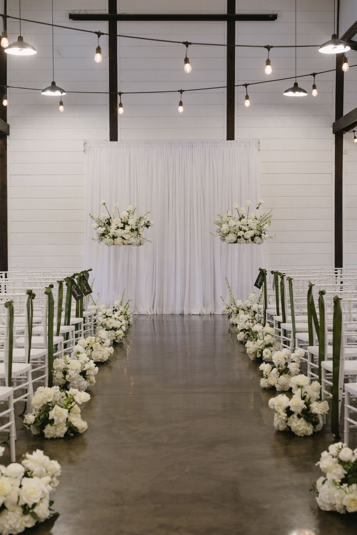 Elegant wedding ceremony space with white floral arrangements and string lights against white brick walls.