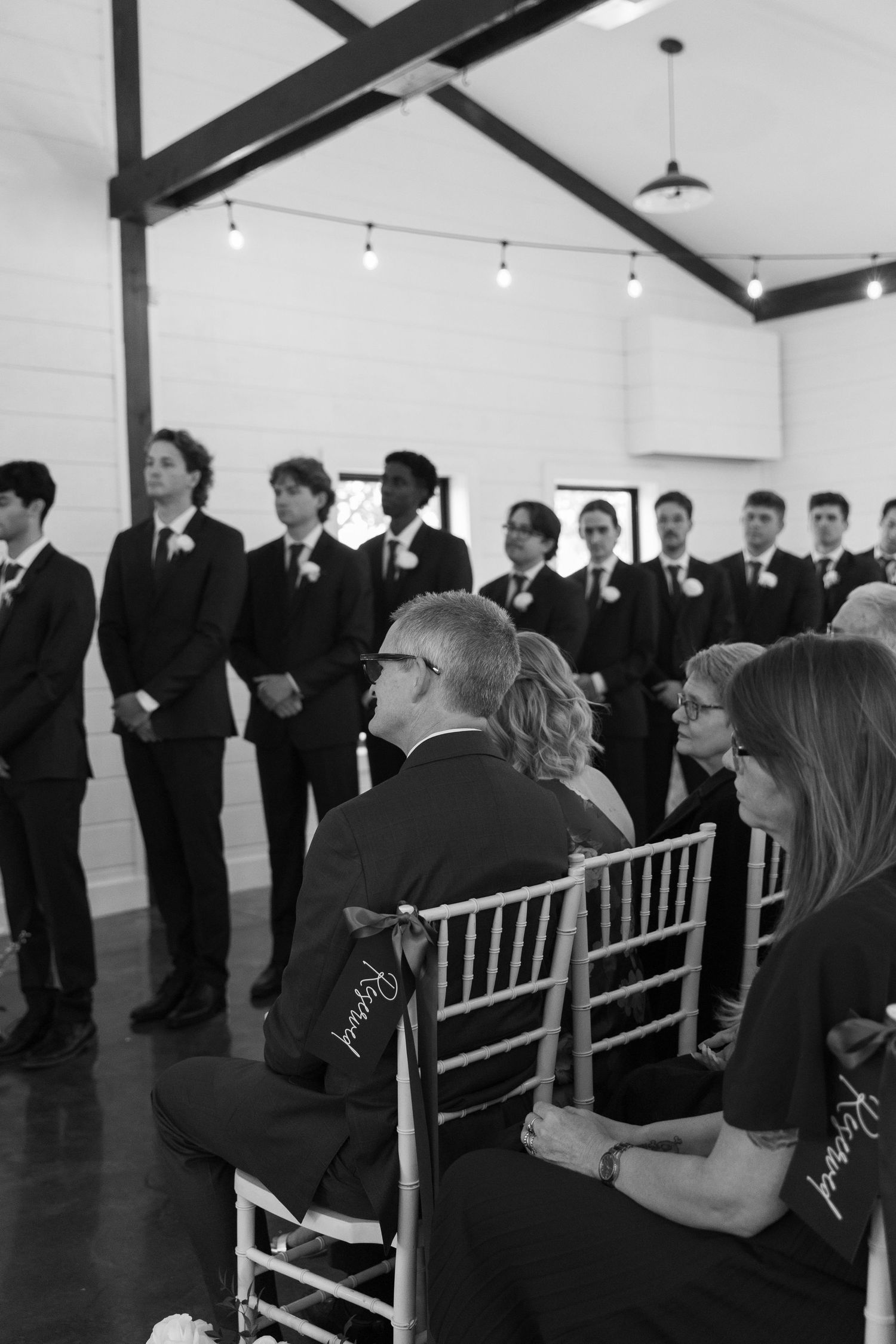 Wedding guests seated in white chairs watch attentively during an indoor ceremony.