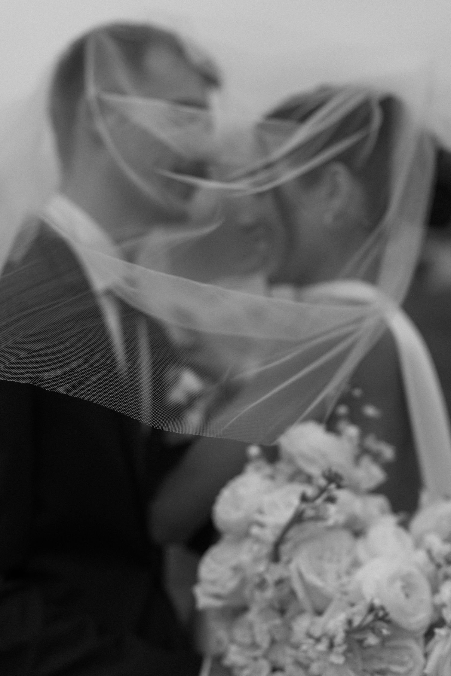 An artistic black and white photo of newlyweds under a flowing veil with white flowers.
