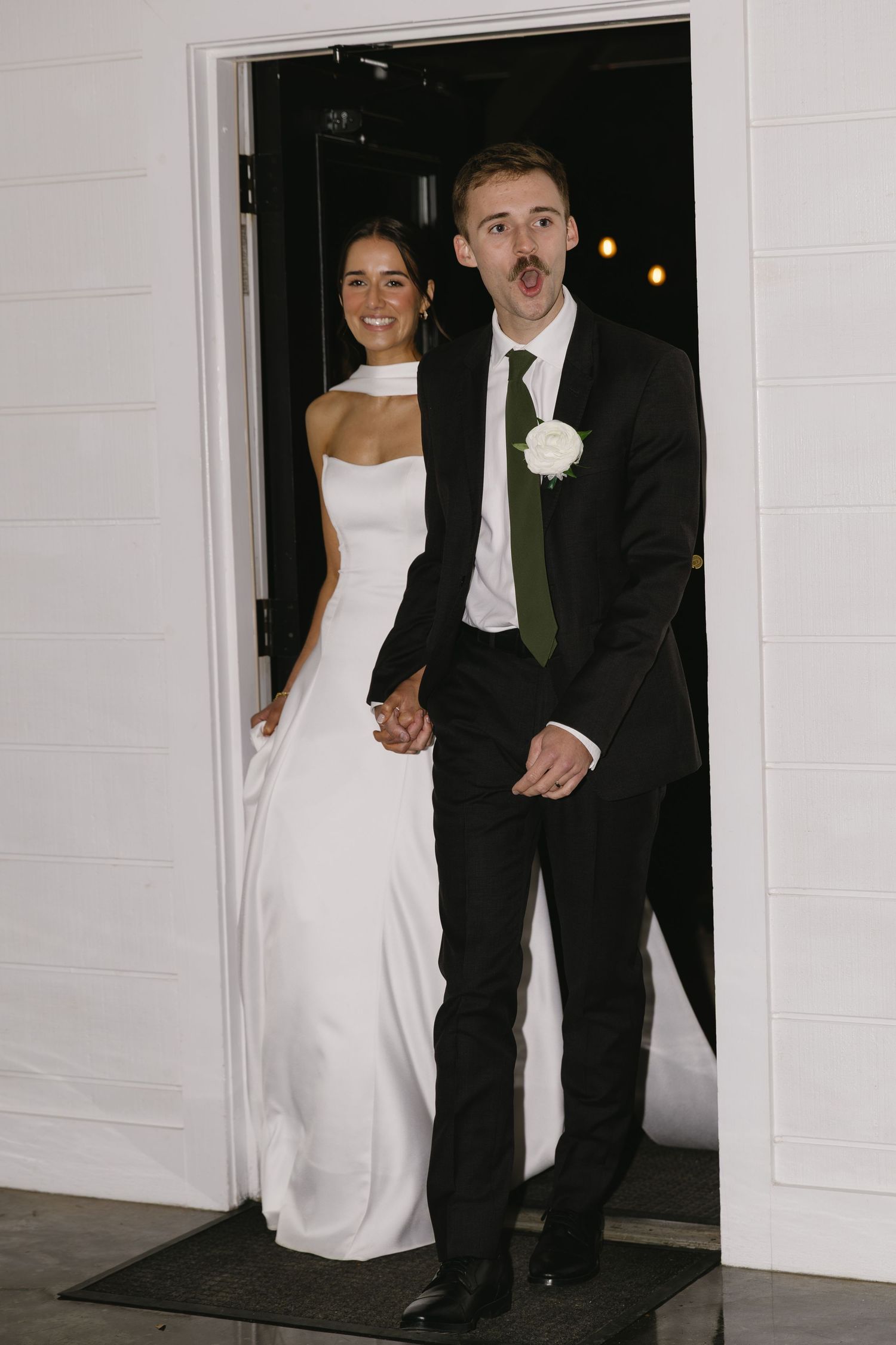 Wedding couple walking out of a white building entrance in formal attire.