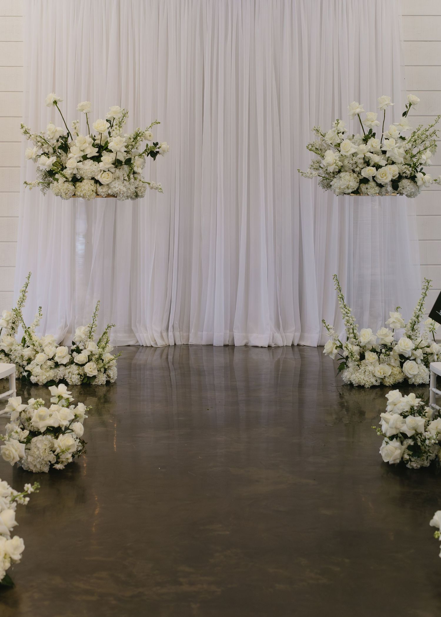 Elegant white wedding ceremony backdrop with floral arrangements and flowing drapes.