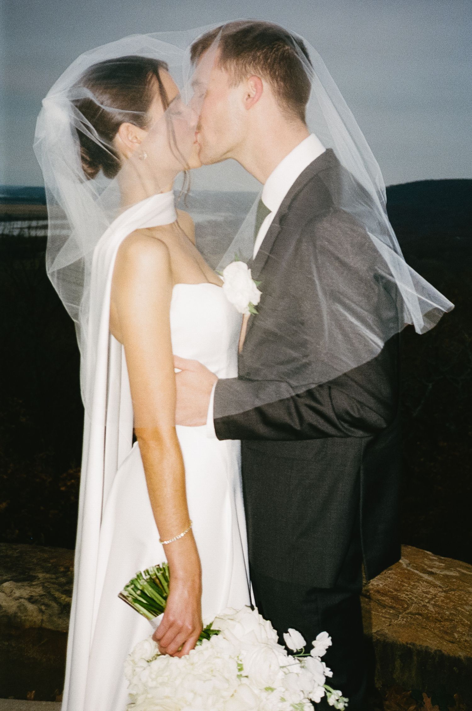 Romantic wedding kiss under veil with bouquet against scenic backdrop.