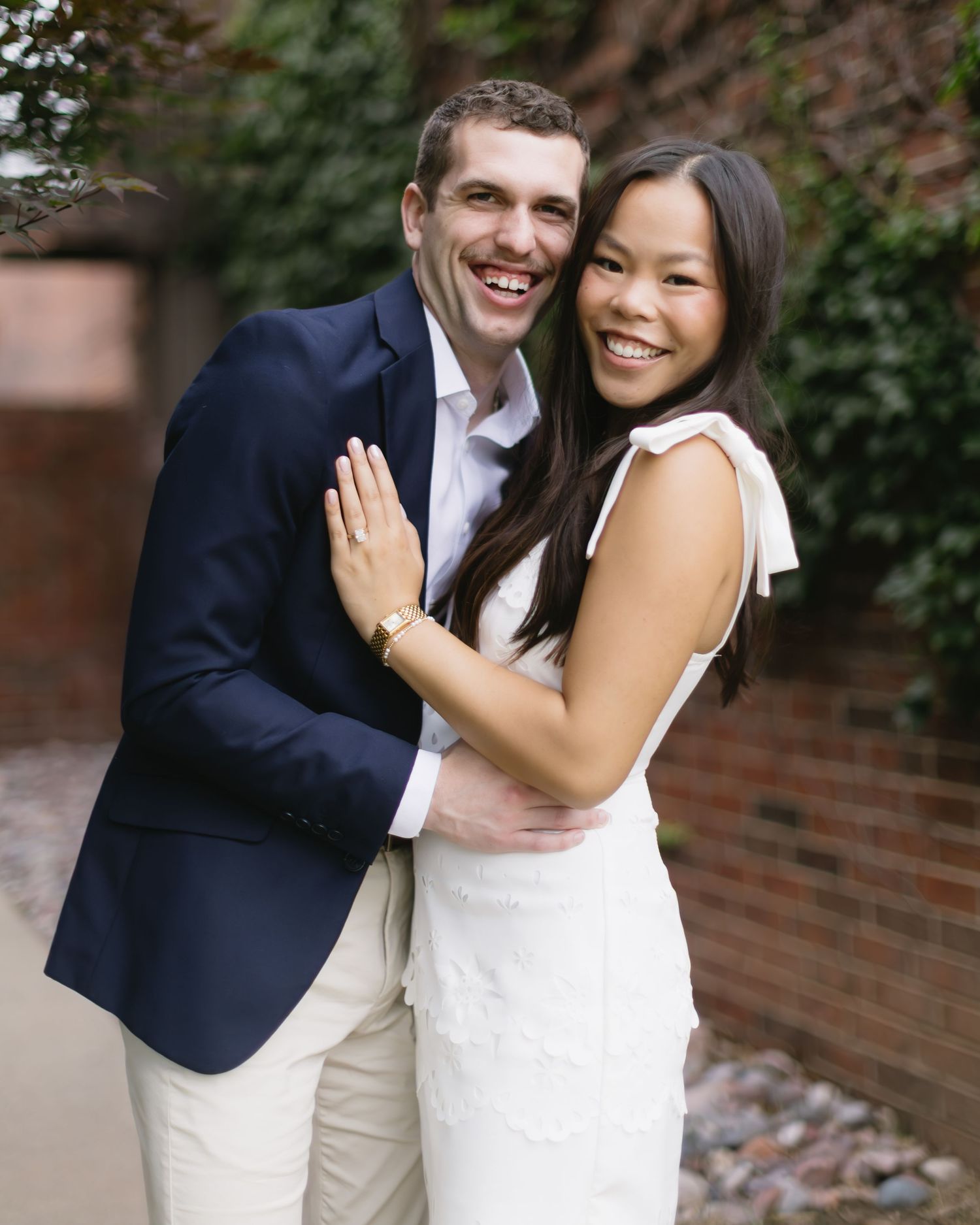 A joyful couple embraces while standing near a brick wall in formal wedding attire.