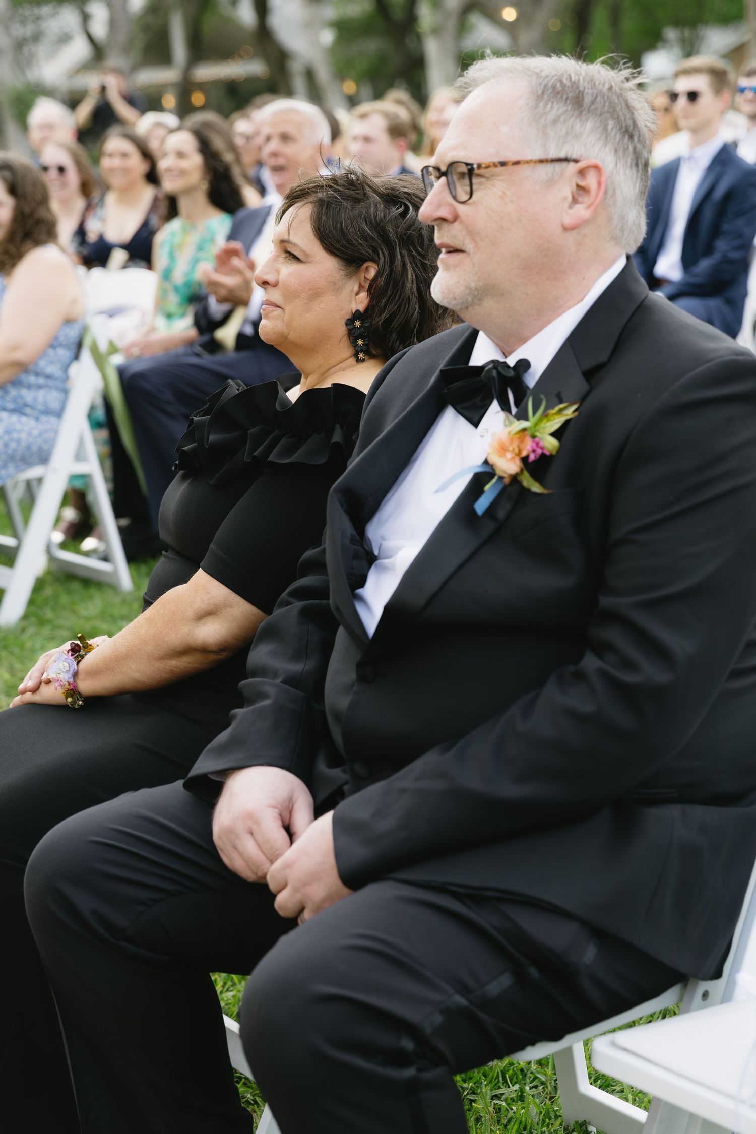 Two guests in formal black attire watching a wedding ceremony from white folding chairs outdoors.