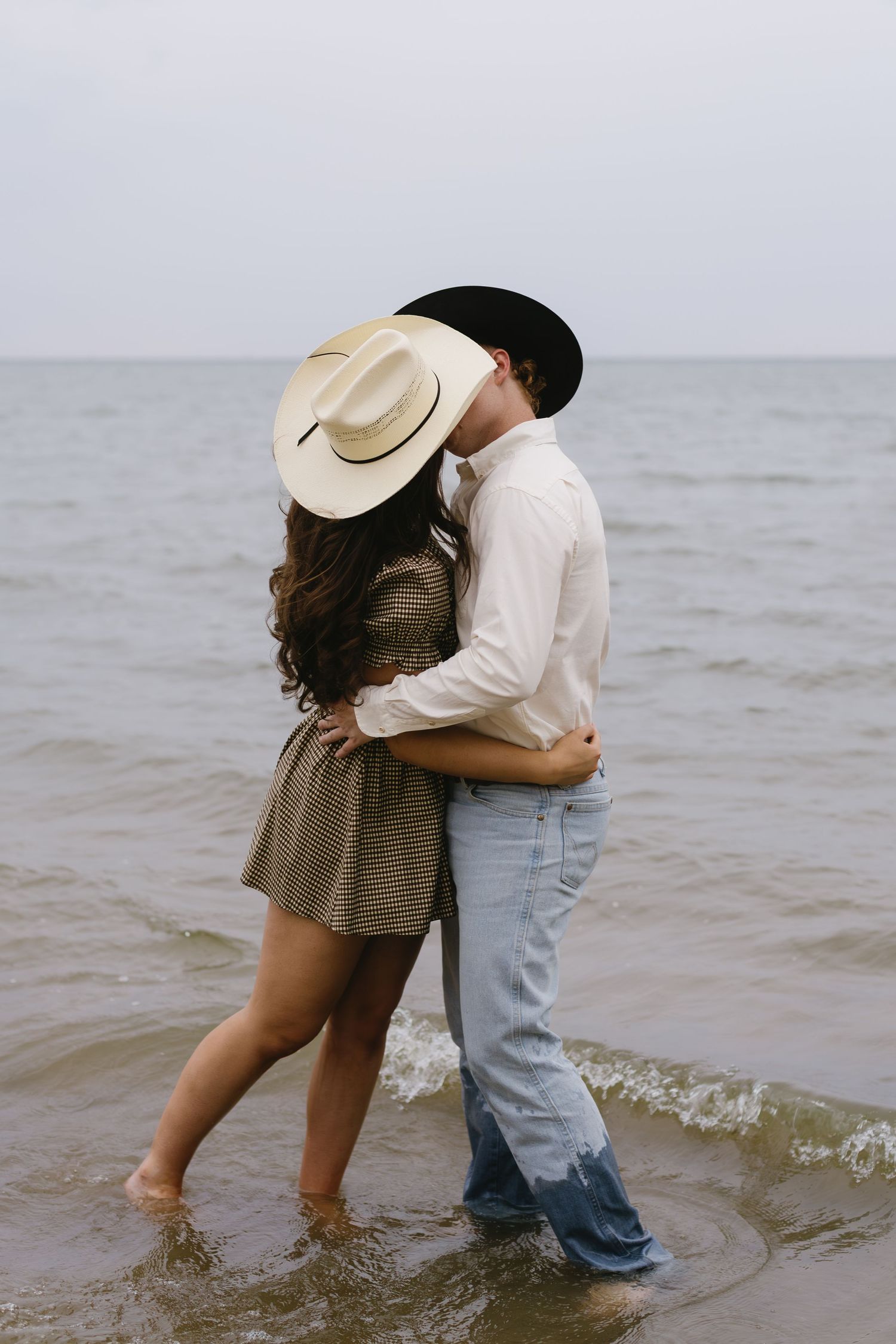 Affectionate couple wading in ocean waves together while wearing western attire and sharing embraces.