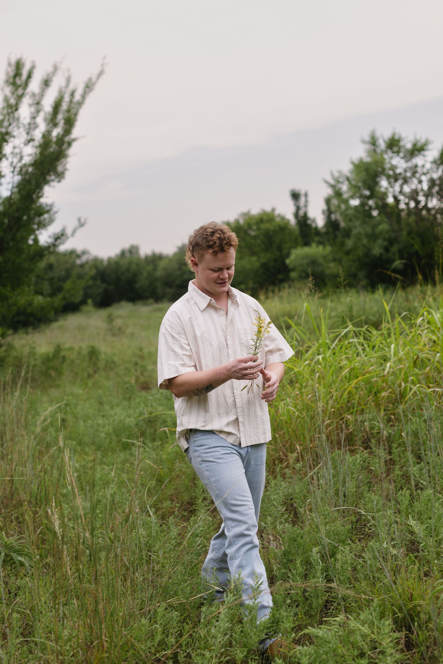 Person in white shirt and jeans walks through tall grass in a countryside field on an overcast day.