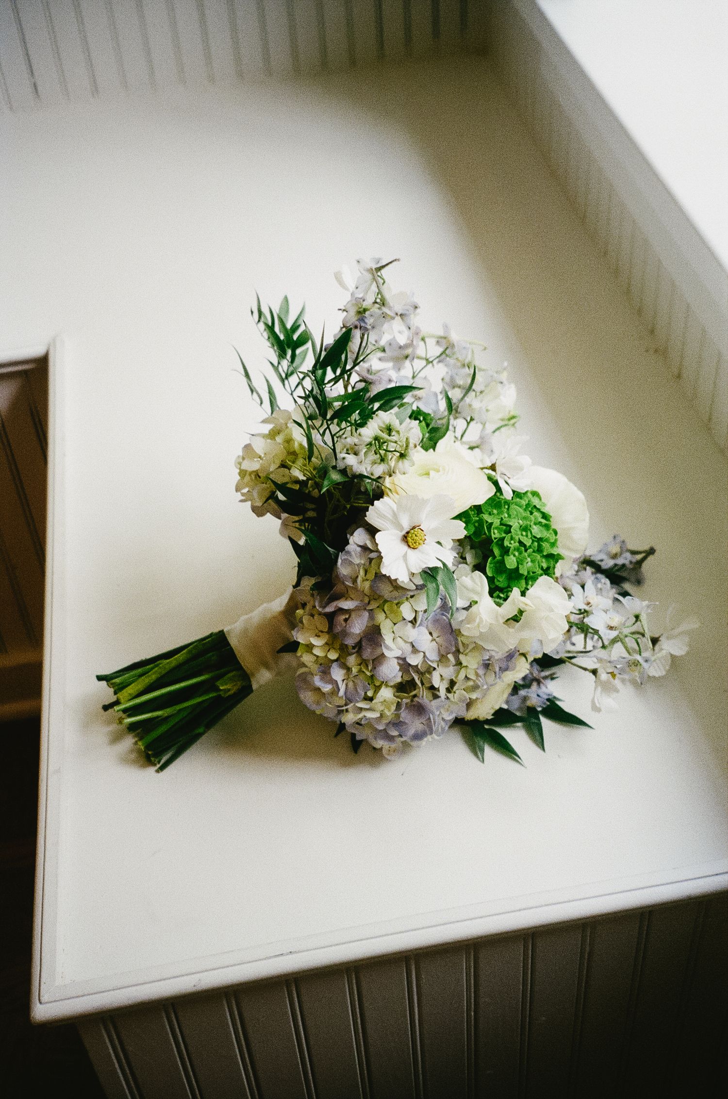 White and purple wedding bouquet with greenery lying on a white surface.