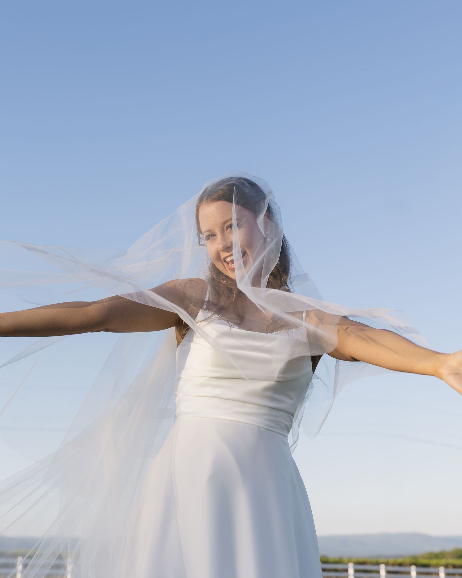 A bride spreads her arms wide, veil flowing in the wind against a bright blue sky.