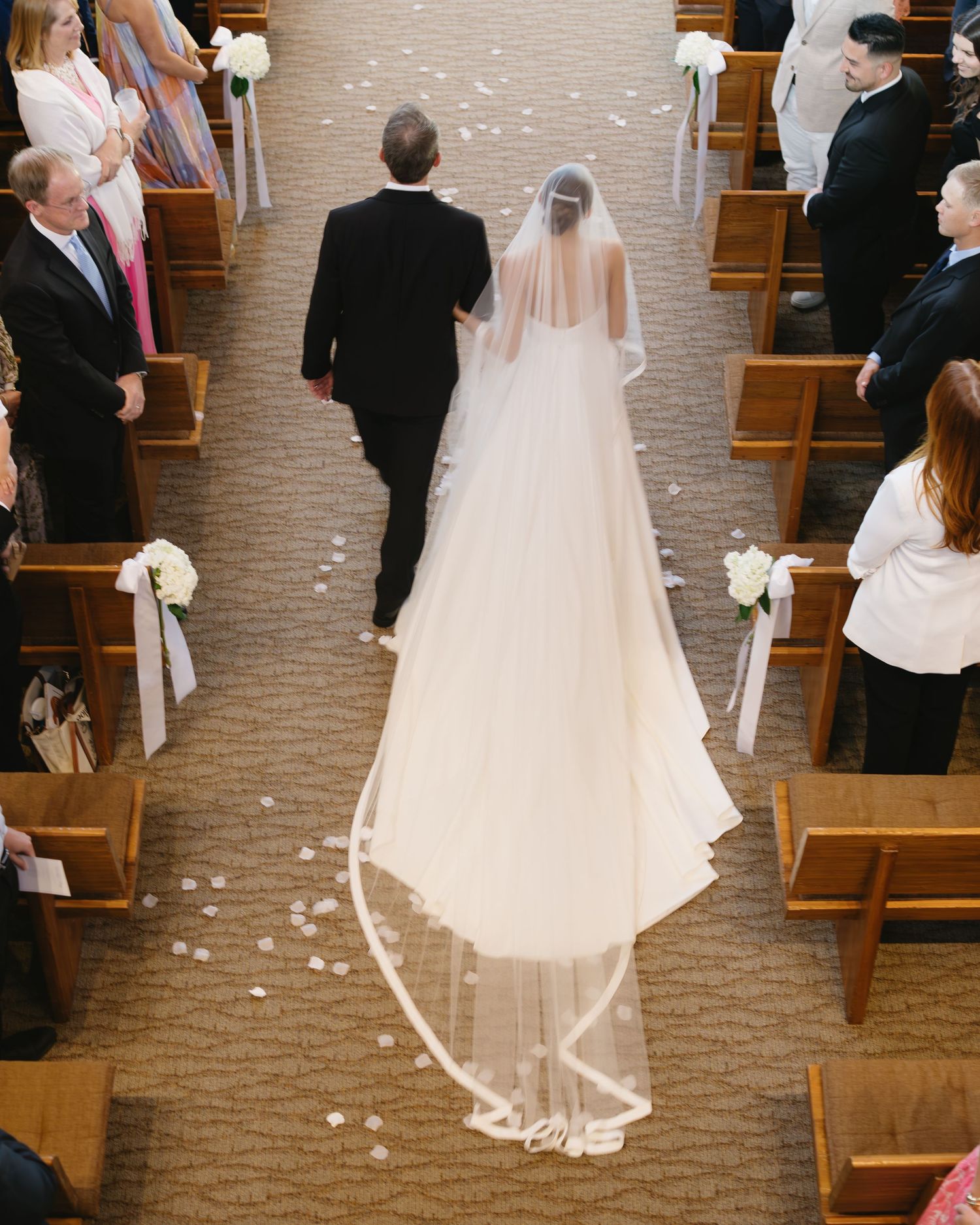 A bride and groom walk down the church aisle with a long flowing veil trailing behind them on a carpet lined with flowers.