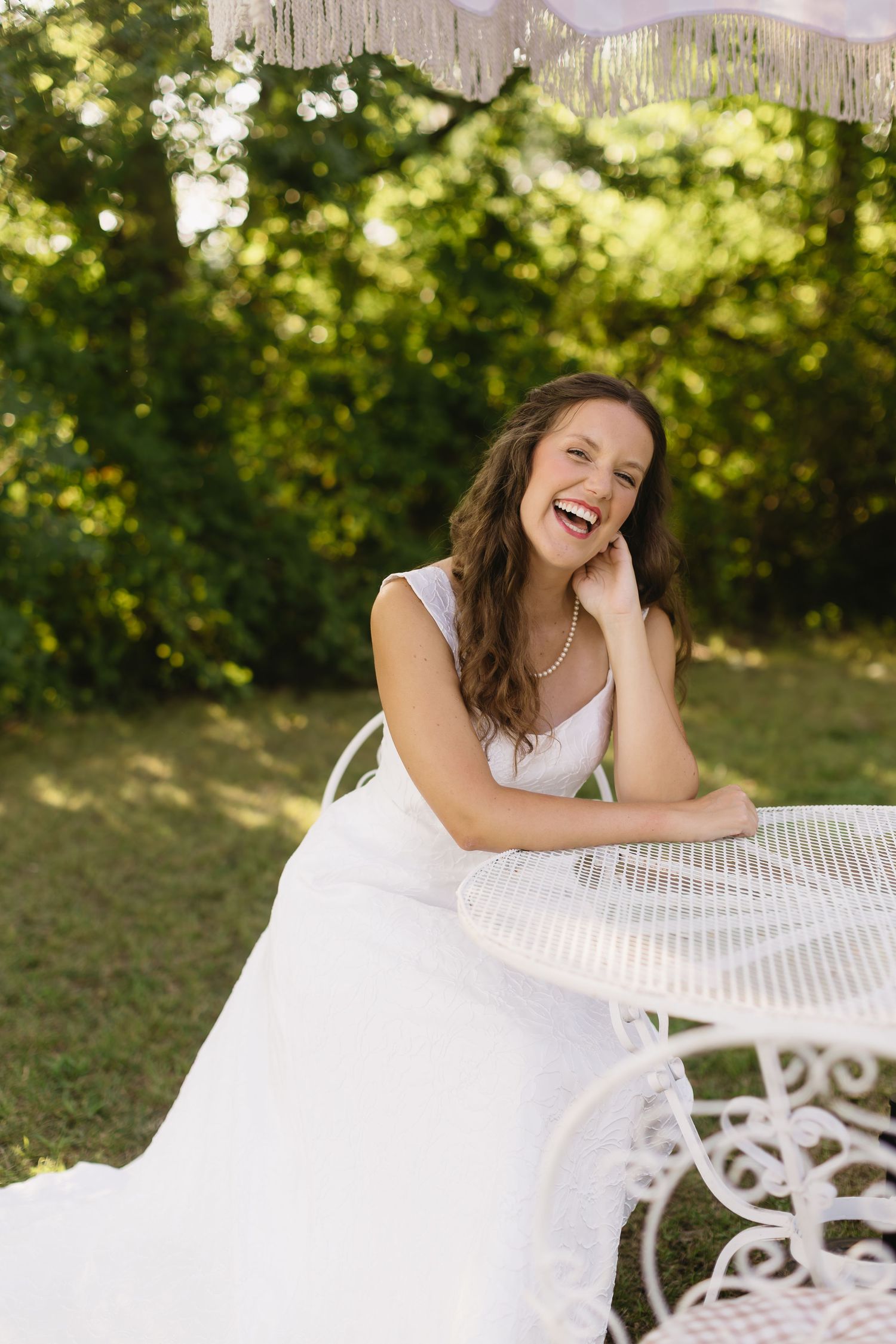 A bride relaxes at a white vintage table set under a decorative parasol in a garden setting.