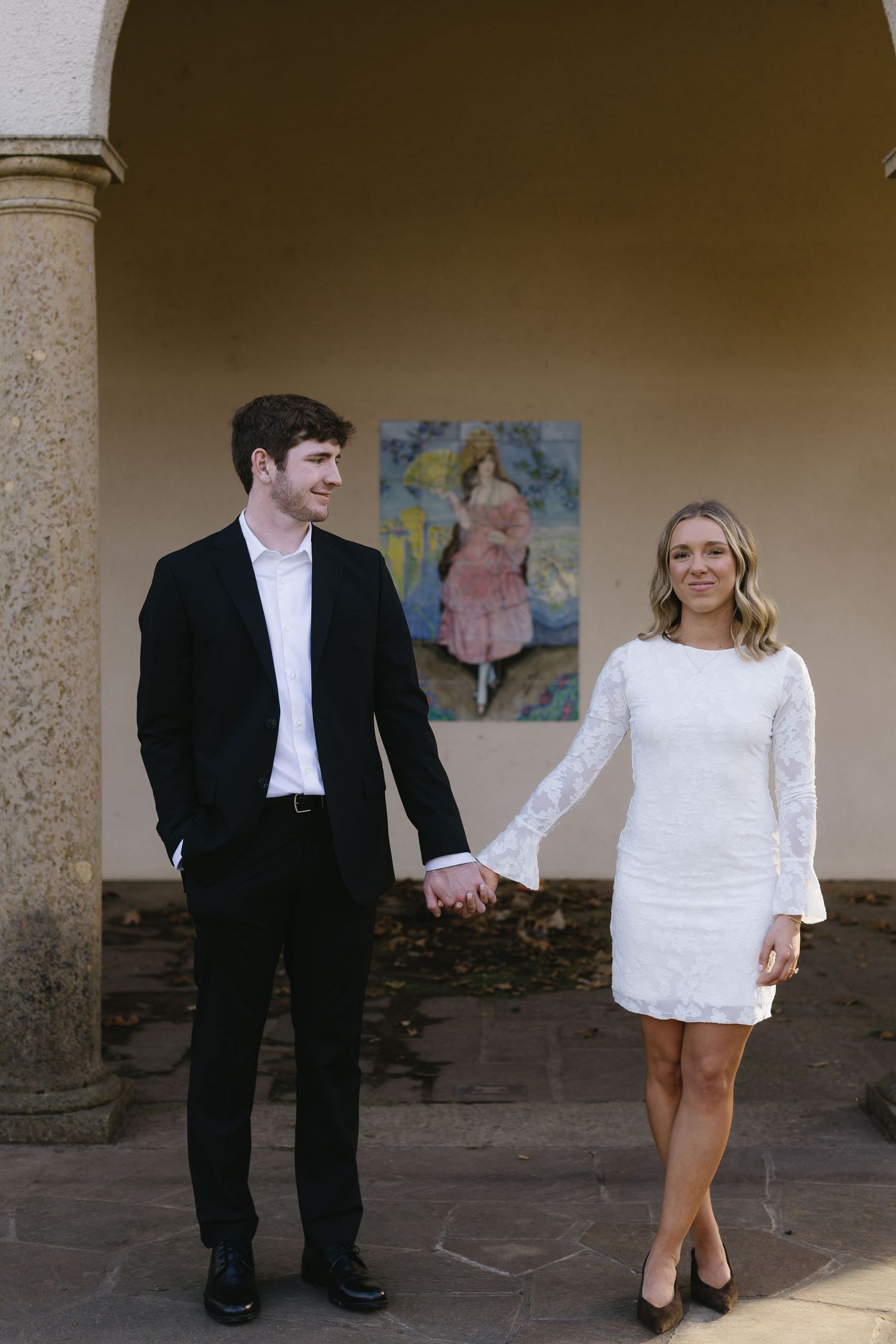 A couple in formal wear holds hands while standing under an archway with artwork.