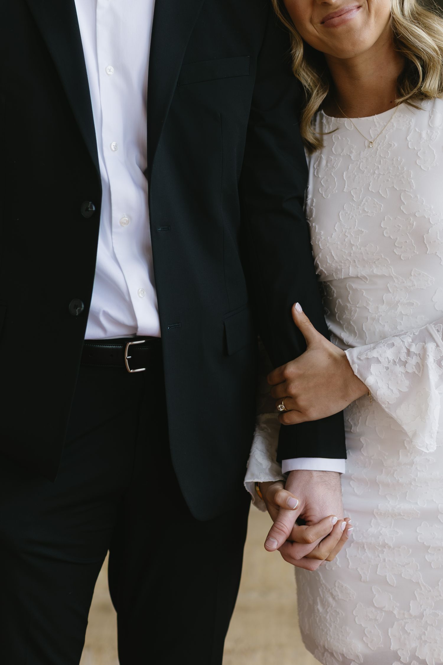 A couple in formal wedding attire embracing, showing close-up details of a black suit and white lace dress.