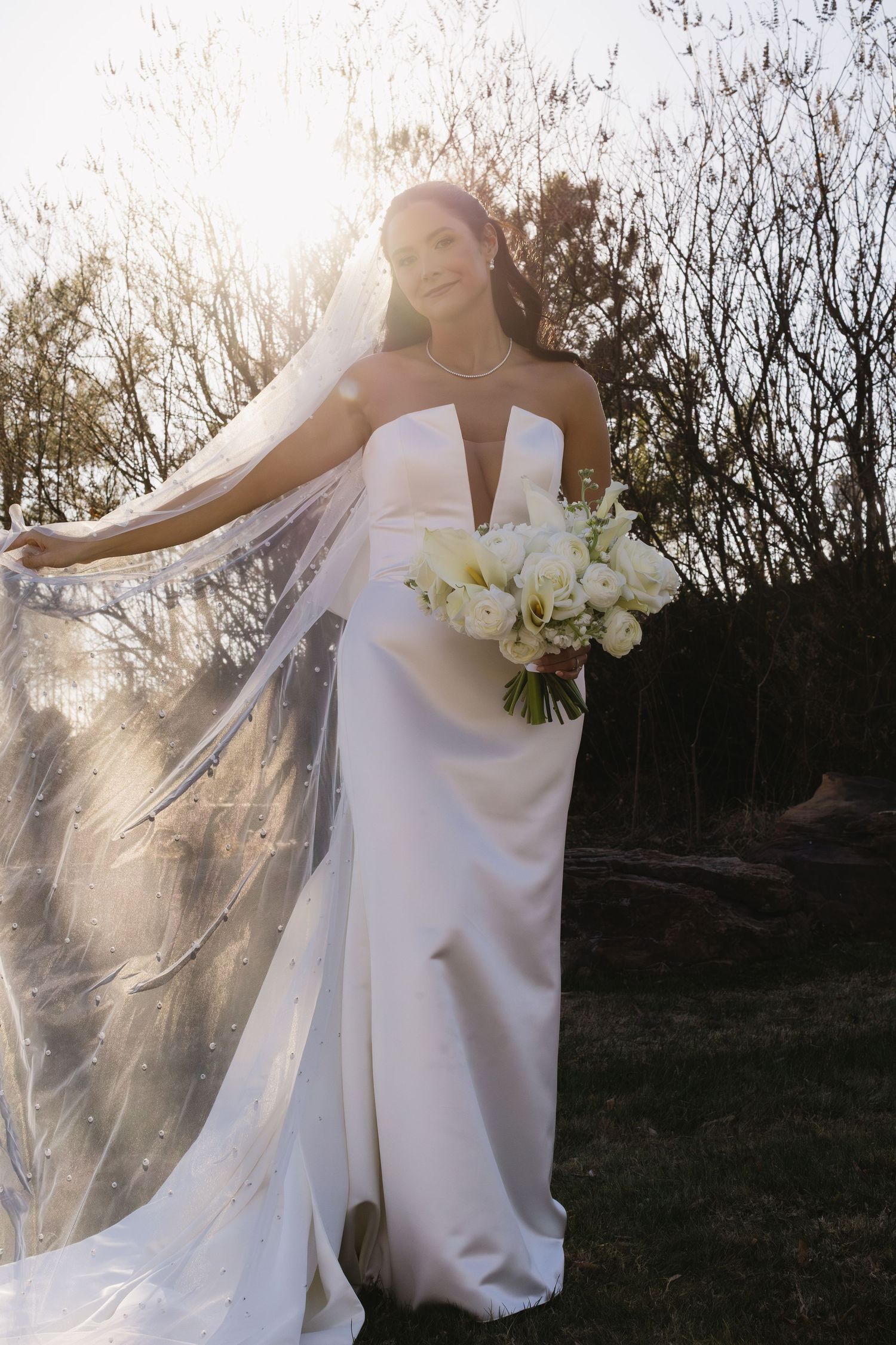 Elegant white wedding dress and veil flowing in the breeze against a natural outdoor backdrop.