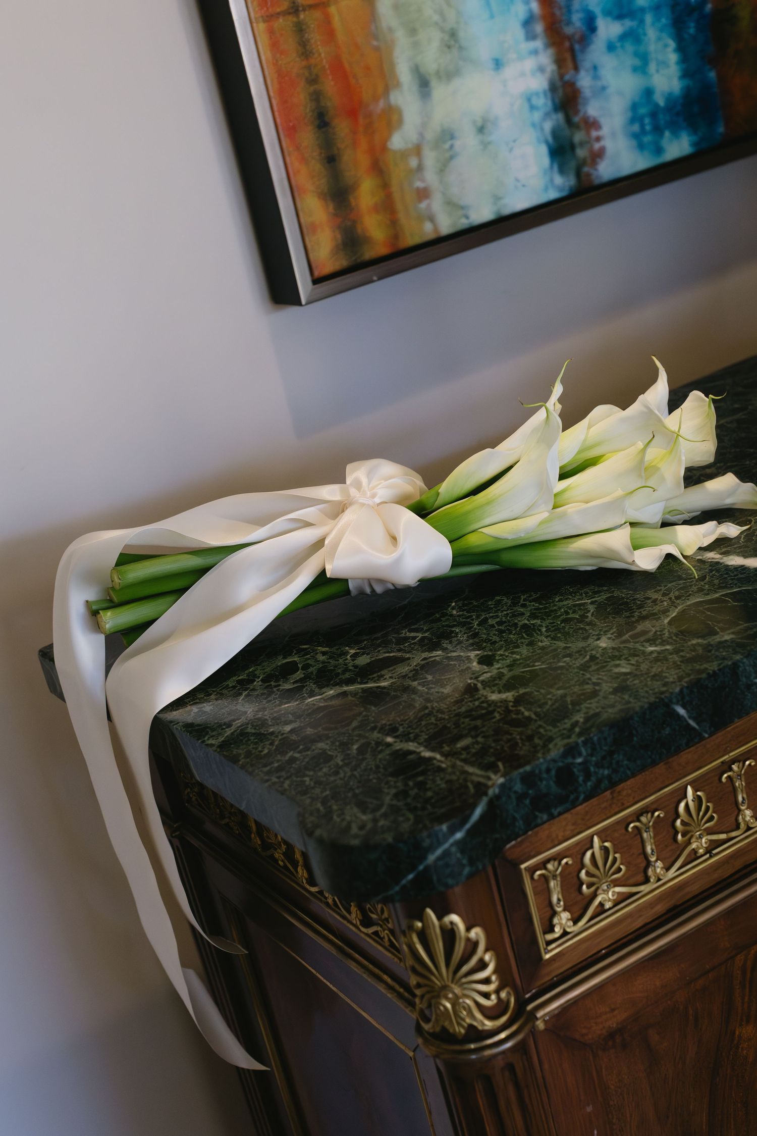 White calla lilies rest on an ornate dark wooden surface with abstract art in background.
