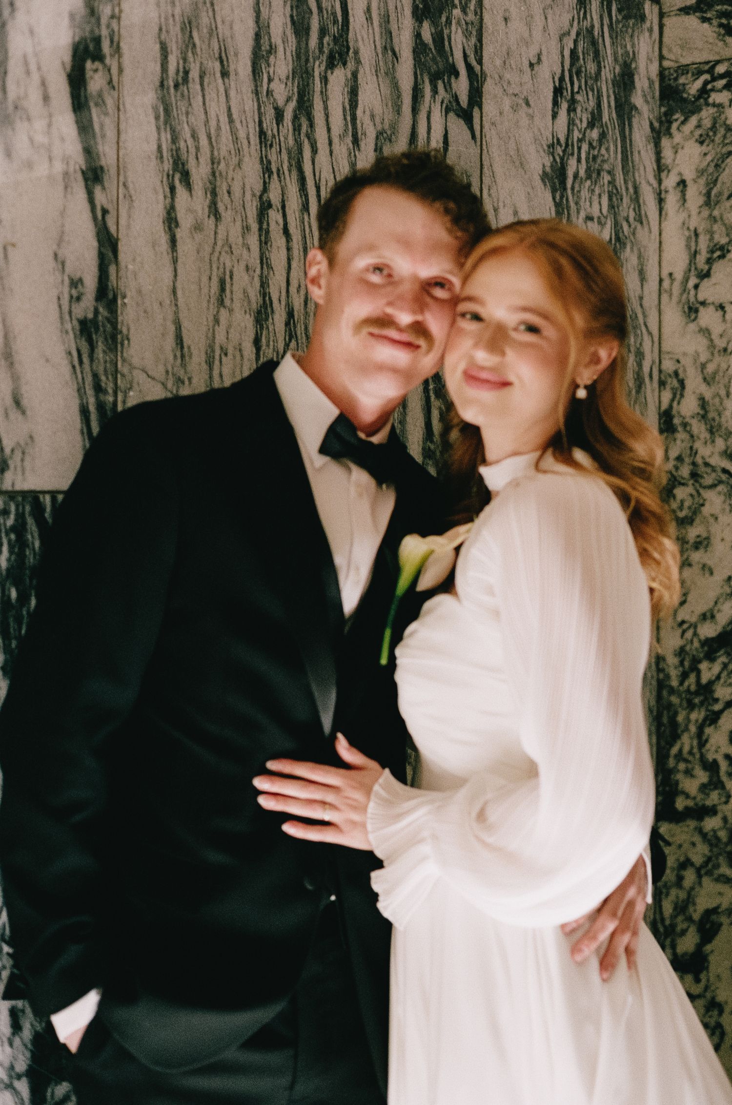 Romantic couple poses against marble wall in wedding attire.