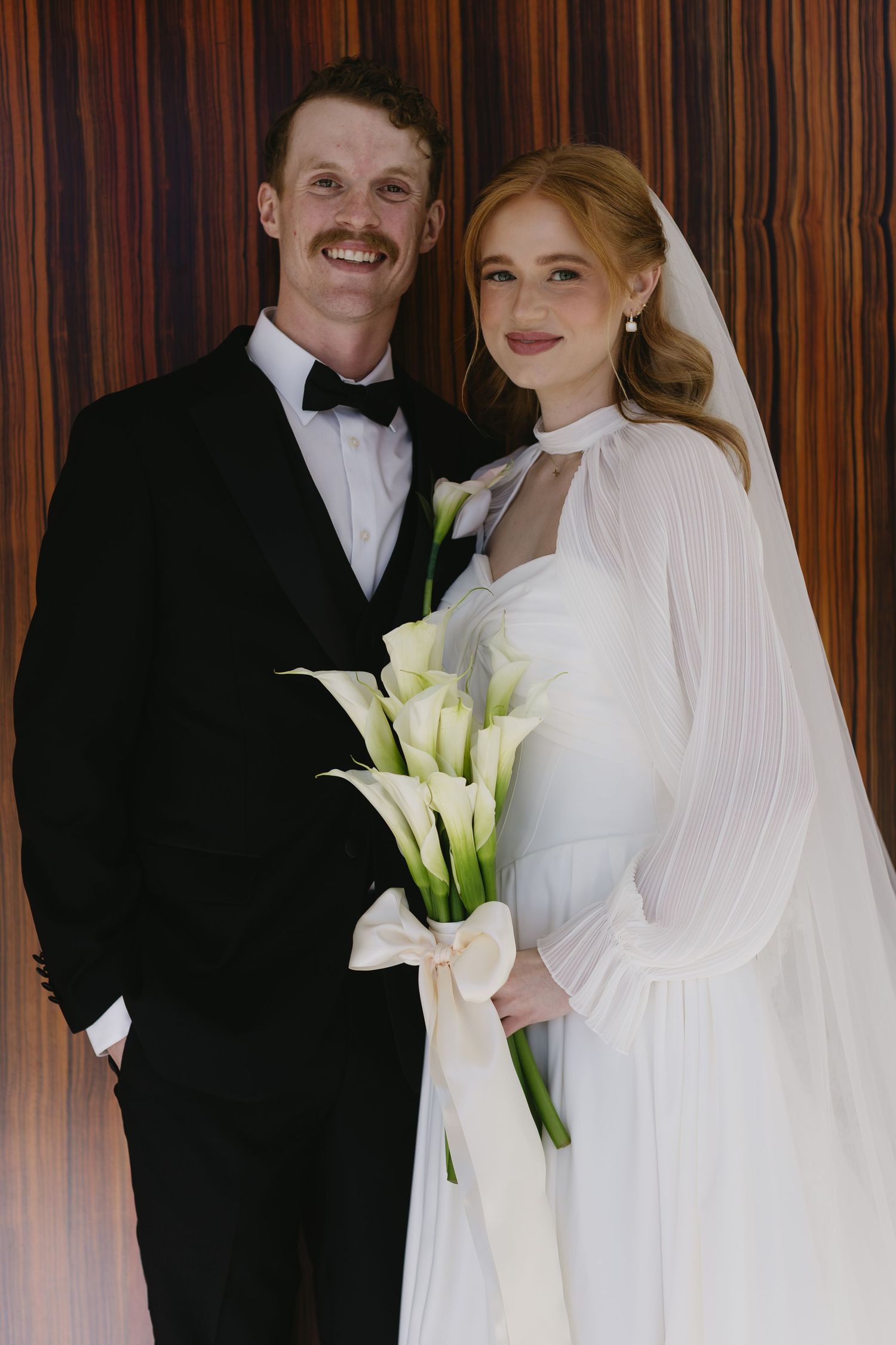 Wedding couple poses together with white calla lily bouquet against wood paneled wall.