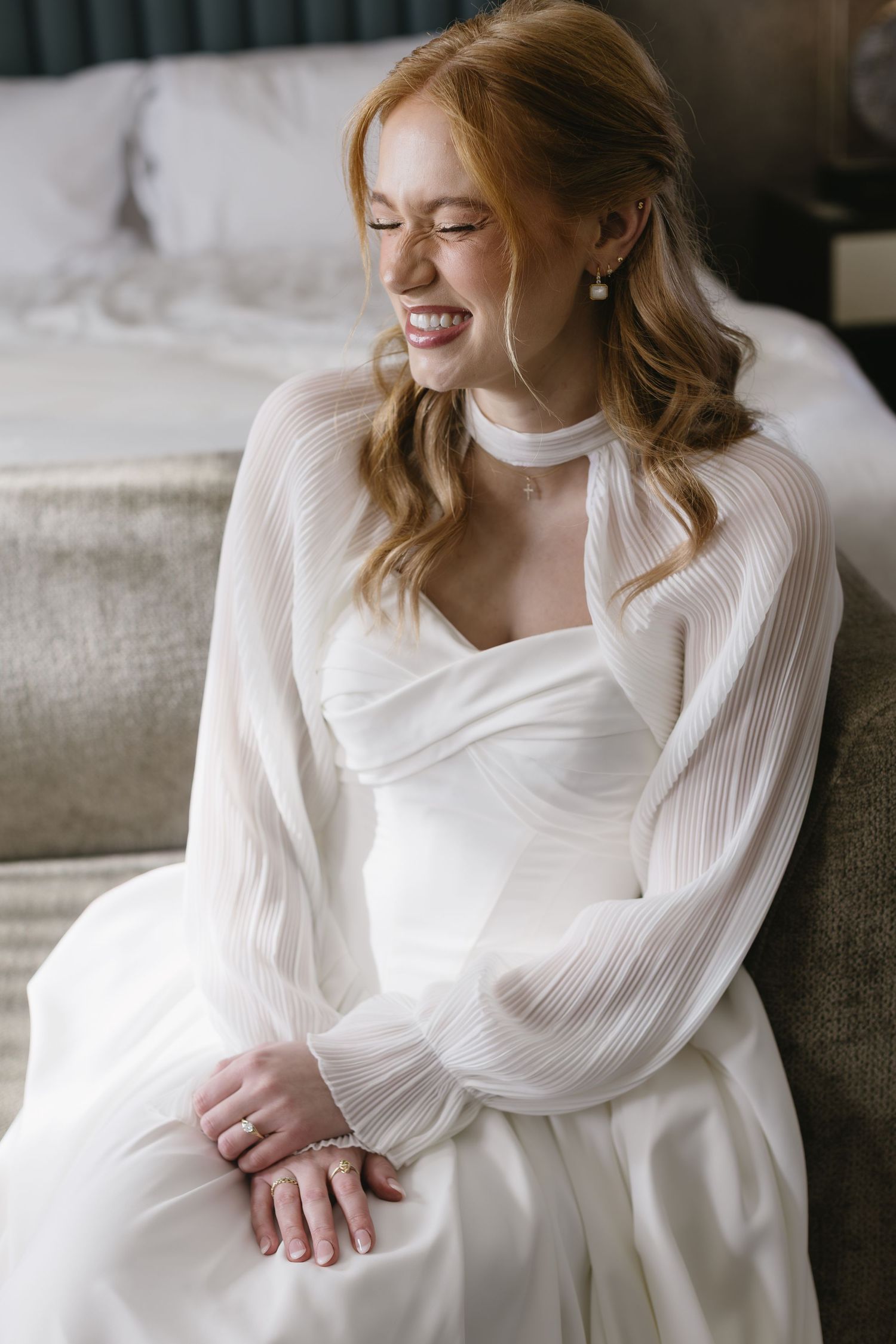 Bride in an elegant white dress with pleated sleeves poses on a couch in natural light.