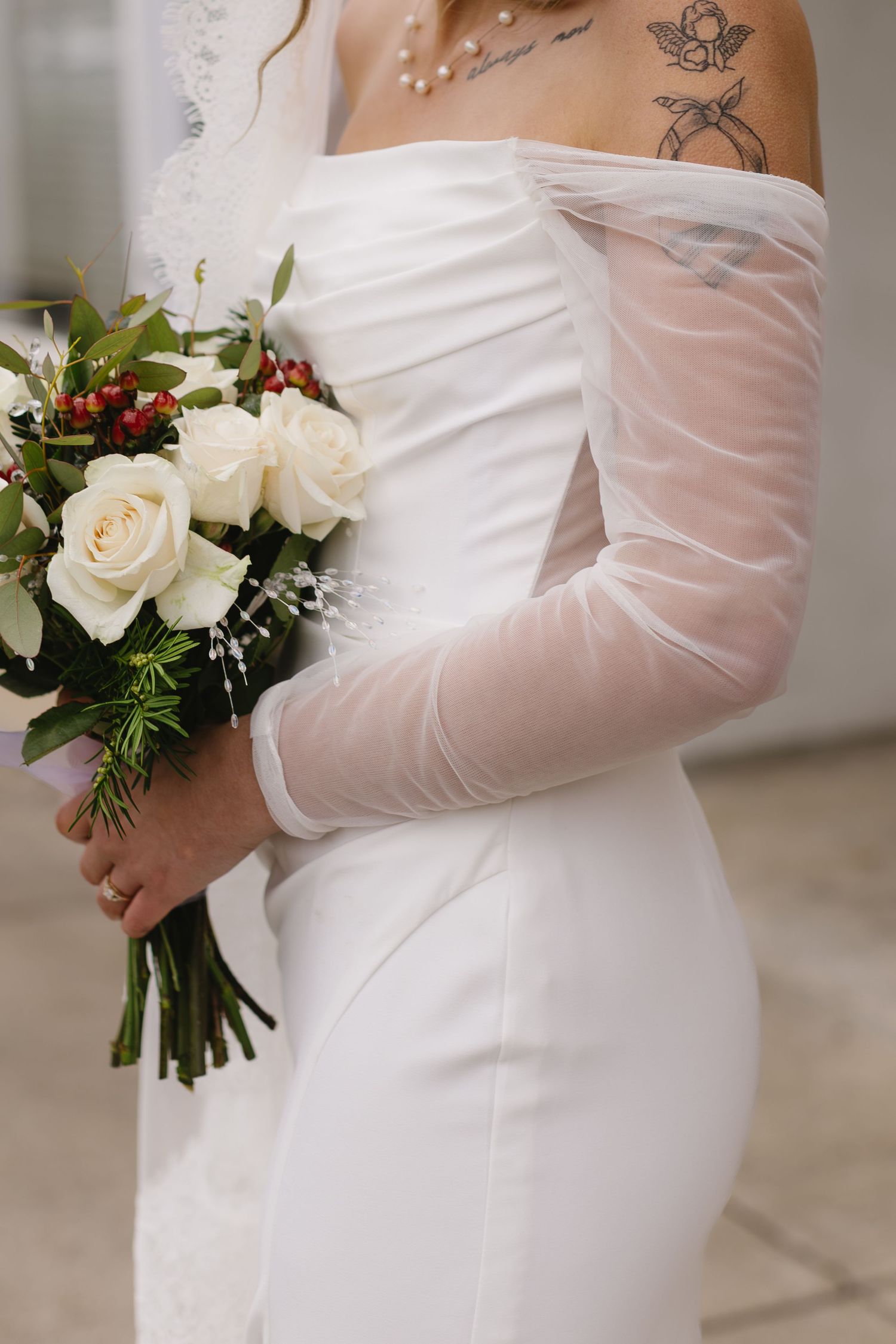 A bride in a modern white gown with sheer sleeves holds a classic white and red winter wedding bouquet.