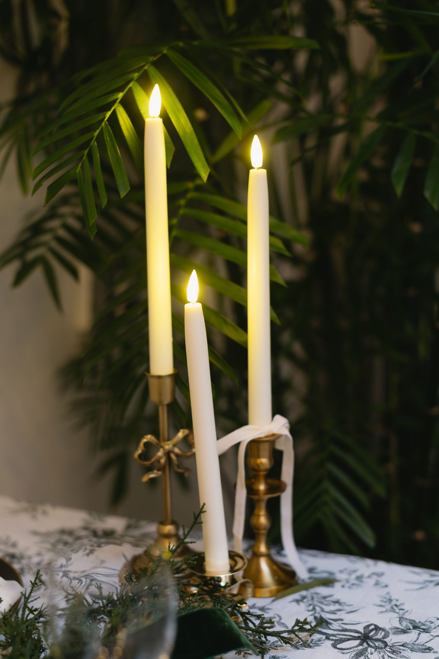 Three lit taper candles in brass holders with tropical foliage backdrop.
