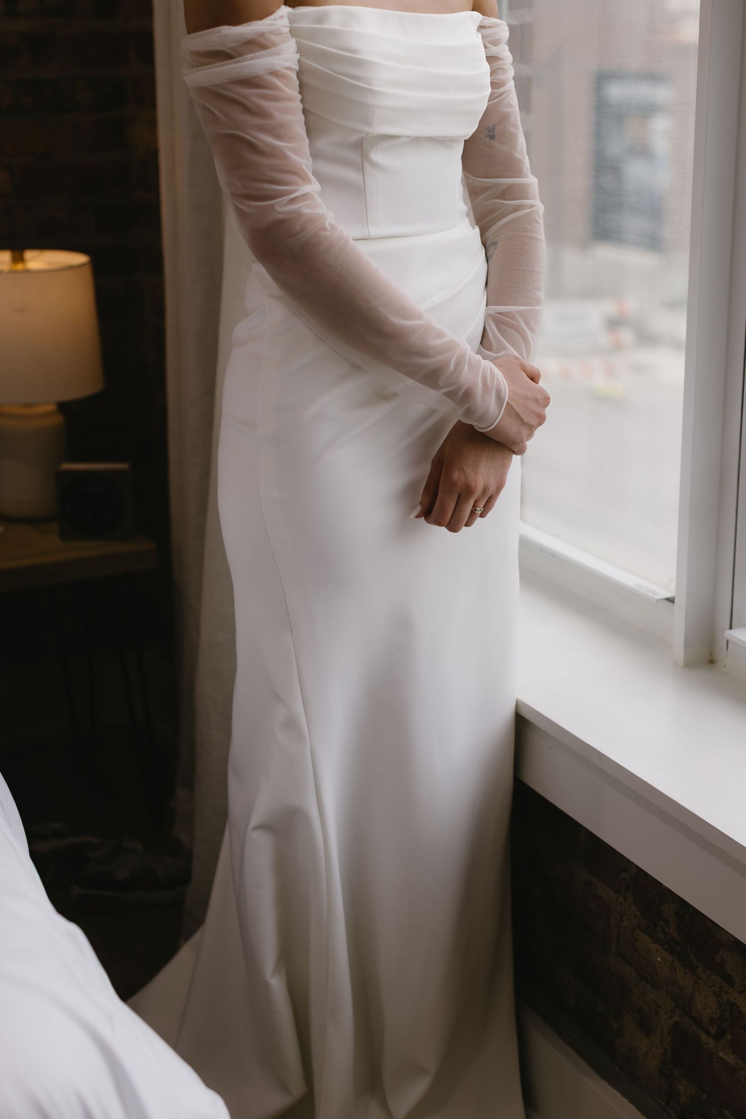 Elegant white wedding dress with sheer long sleeves and fitted silhouette shown near a window.