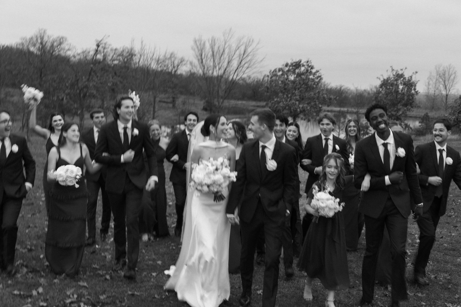 A wedding party walks together outdoors in a black and white photograph with bare trees in the background.